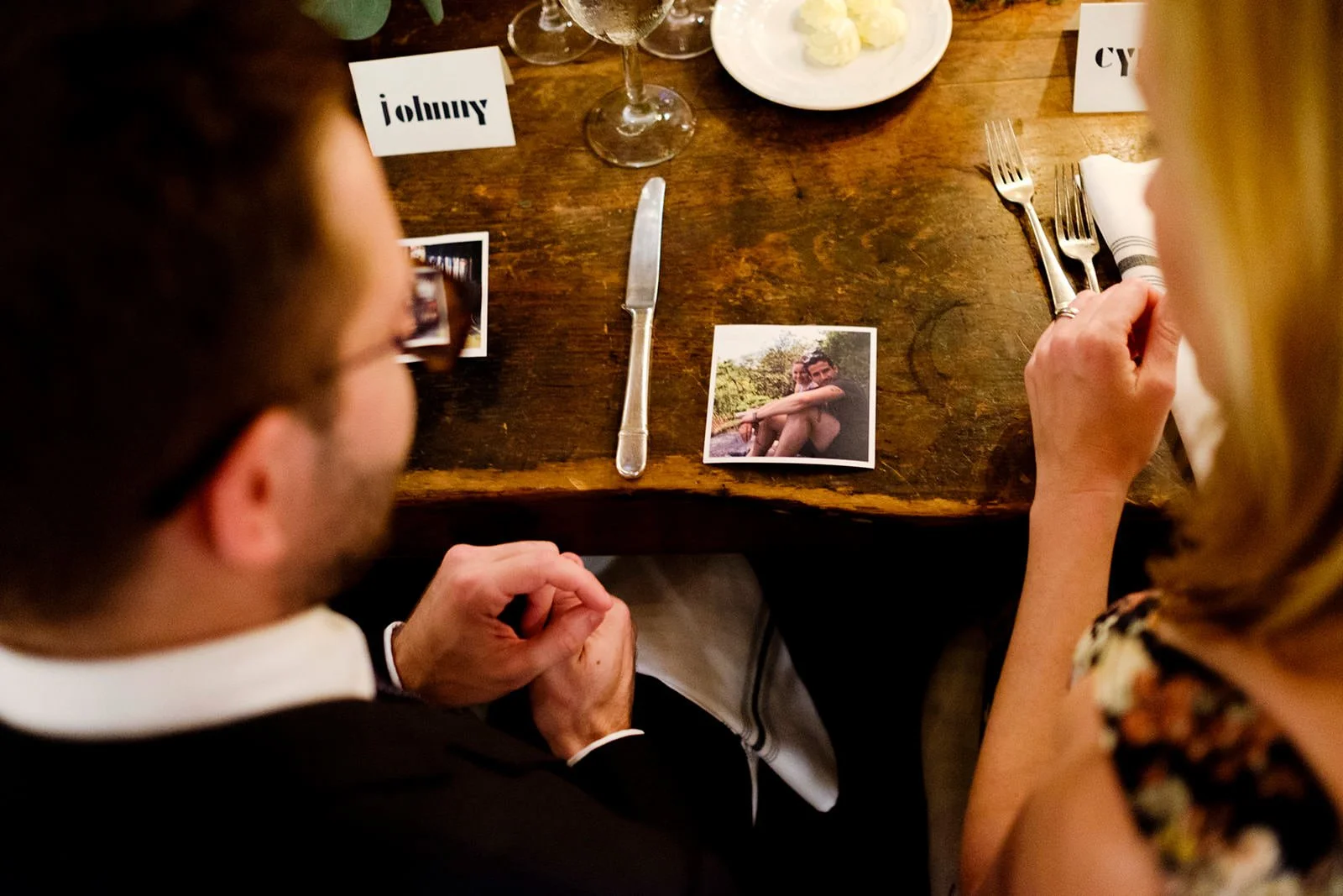 A wooden dining table set for a meal with cutlery, wine glasses, and a plate of food. There are two people's hands visible, one man and one woman, with photographs and name tags on the table.
