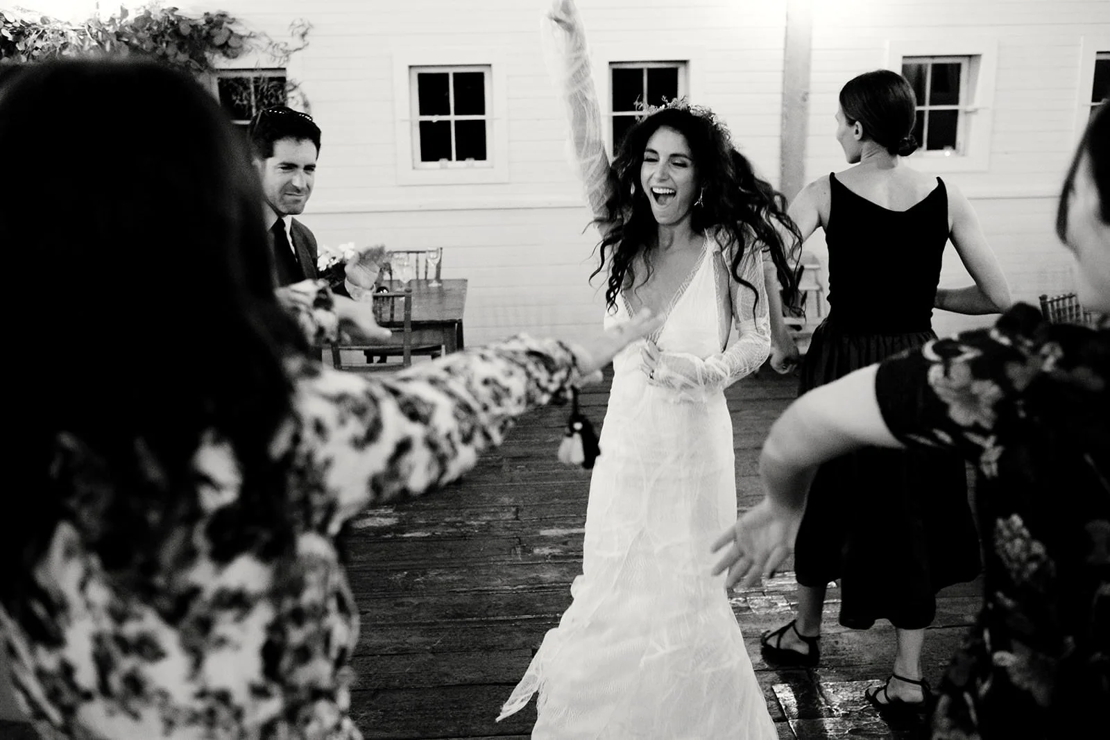 A woman in a white wedding dress dancing and smiling at a party, with other guests around her, in a rustic indoor setting.
