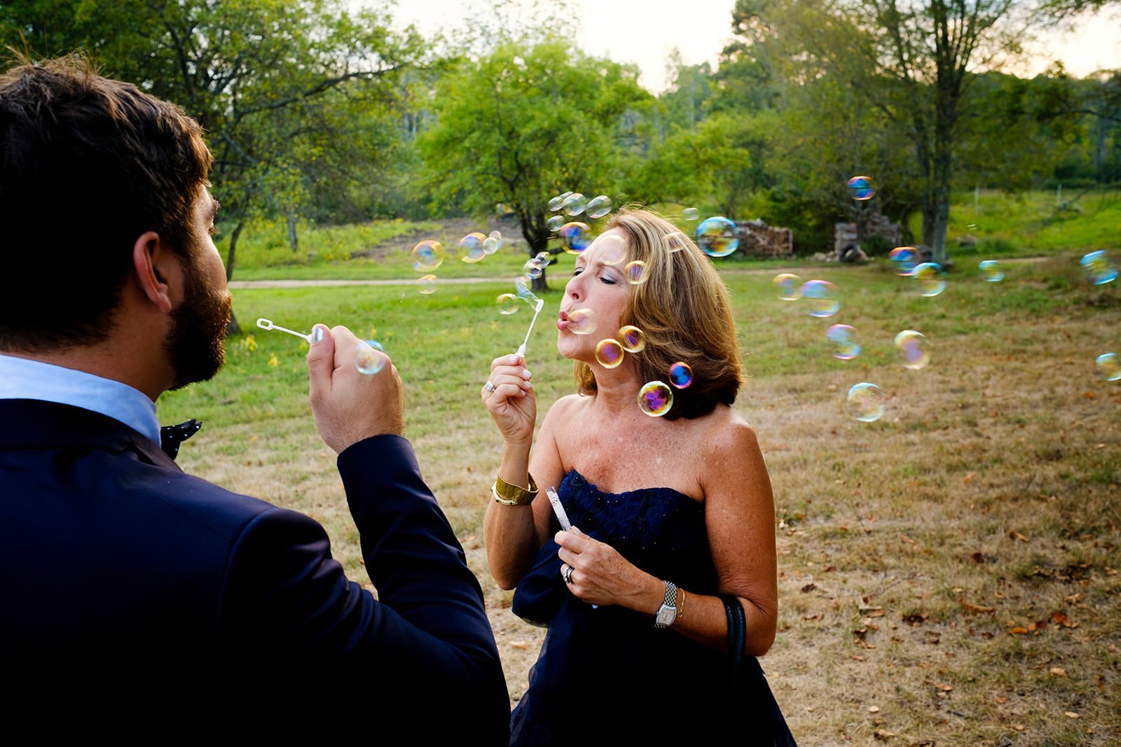 A man and woman blowing bubbles outdoors on a grassy area surrounded by trees.