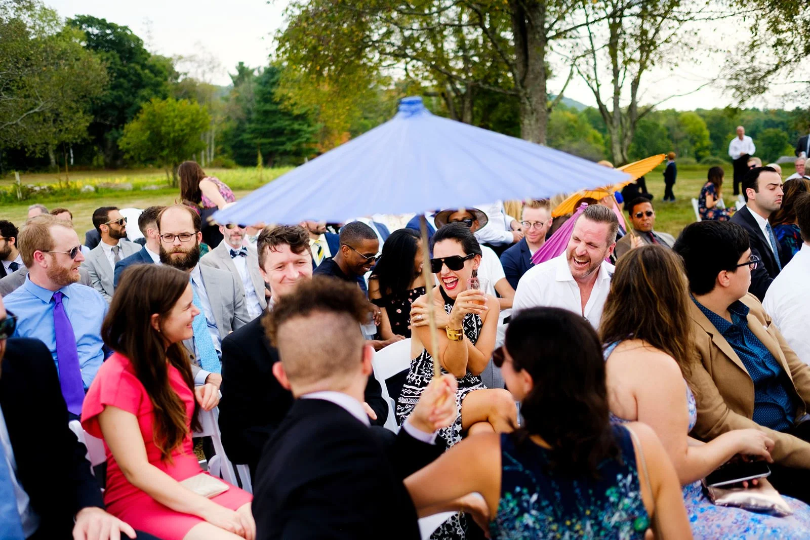 Group of people seated outdoors at a formal event, with a large blue patio umbrella and trees in the background. Some are smiling and laughing, with a woman in black sunglasses holding a drink.