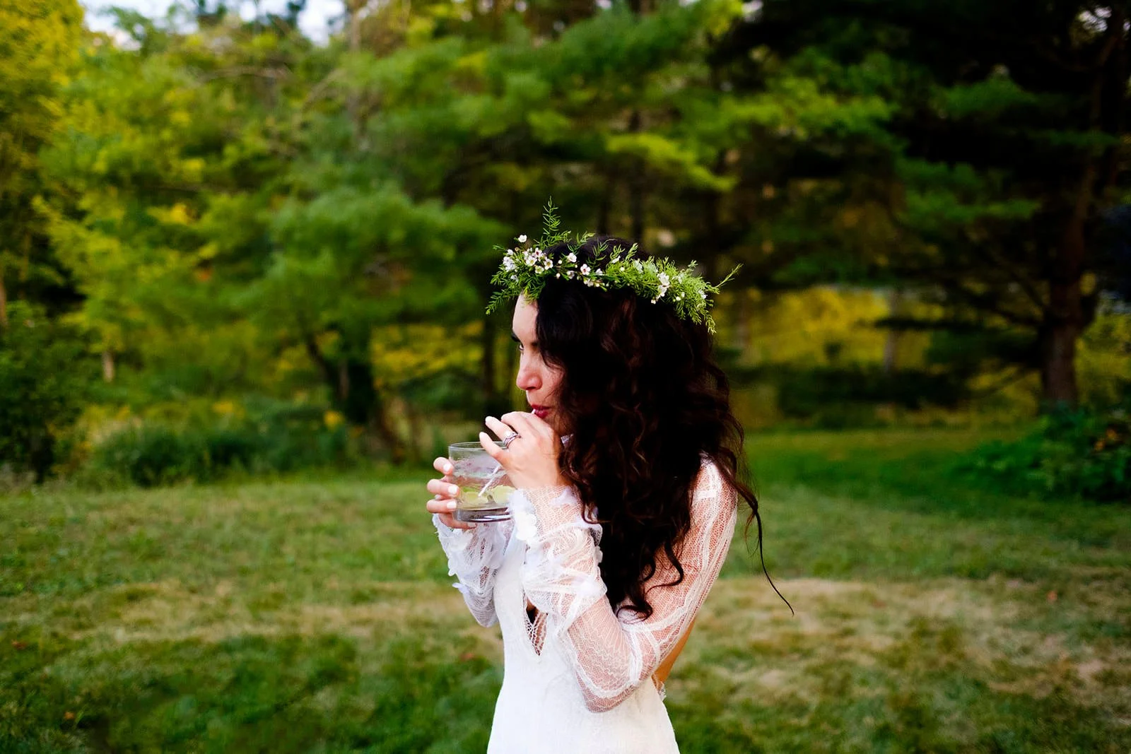 A woman with long dark curly hair wearing a lace white dress and a flower crown, drinking from a glass outdoors in a green park or garden.