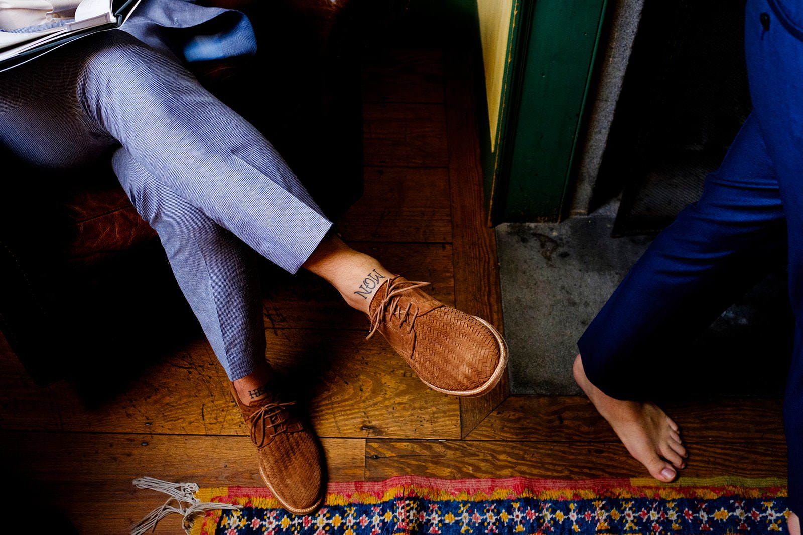 Two people in a room, one seated on a brown leather chair with legs extended, wearing gray checkered pants and brown woven shoes, with tattoos on their ankles. The other person is standing barefoot on a wooden floor next to a colorful rug, wearing da