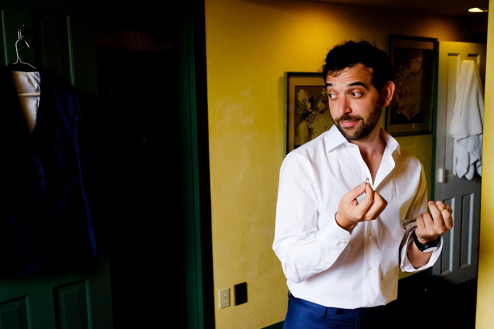 A man in a white dress shirt with cufflinks looks to the side while holding his shirt collar with one hand and a ring with the other, standing inside a room with yellow walls and framed artwork.