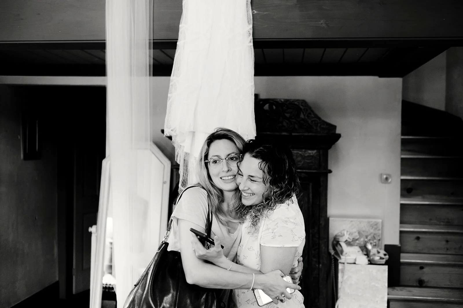 Two women hugging each other indoors, smiling and appears happy, one wearing glasses, the background includes a staircase, a fireplace, and some household items.