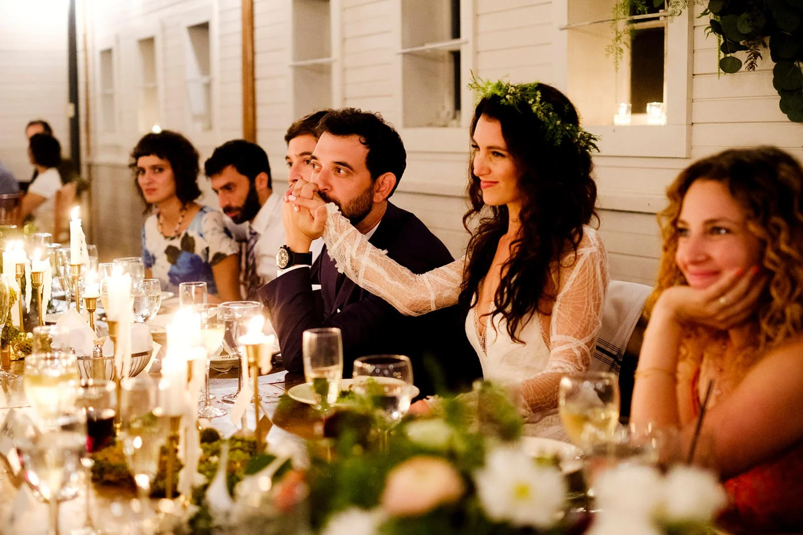 People sitting at a long dinner table during a celebration, with candles and floral decorations, inside a well-lit room with white wooden walls and windows.