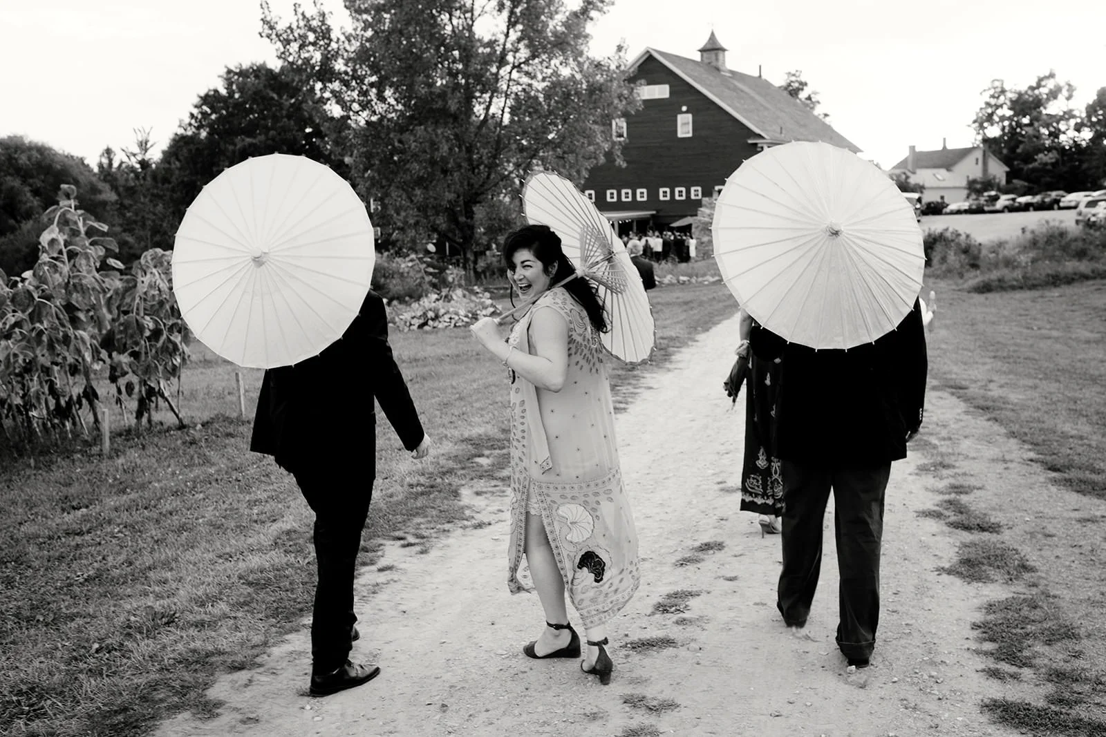 A woman in a patterned dress smiling and holding an umbrella walking on a dirt path, flanked by two men in suits with umbrellas. There is a rustic building and trees in the background.