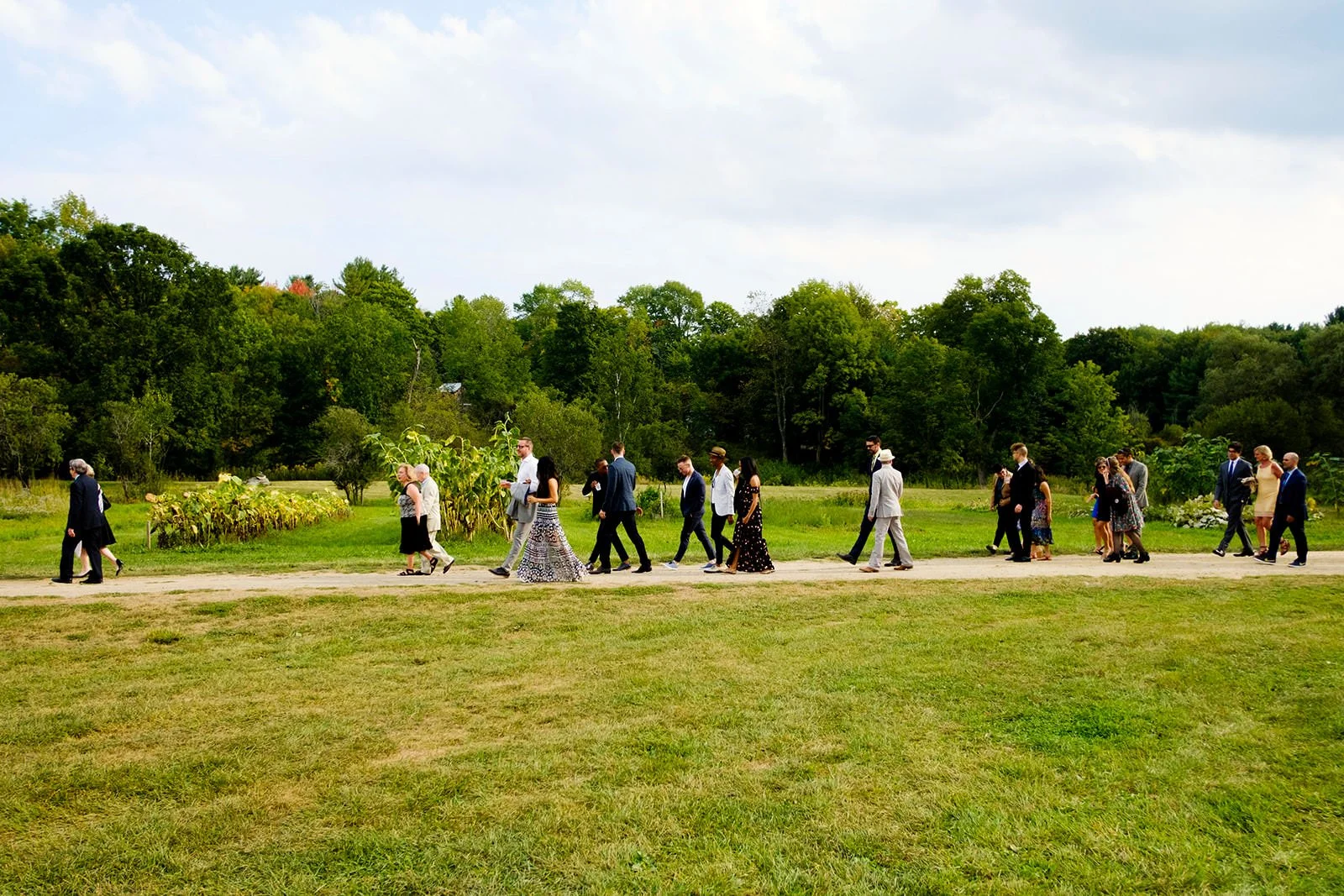 A group of people dressed in formal clothing walking on a dirt path through a grassy field with trees in the background.