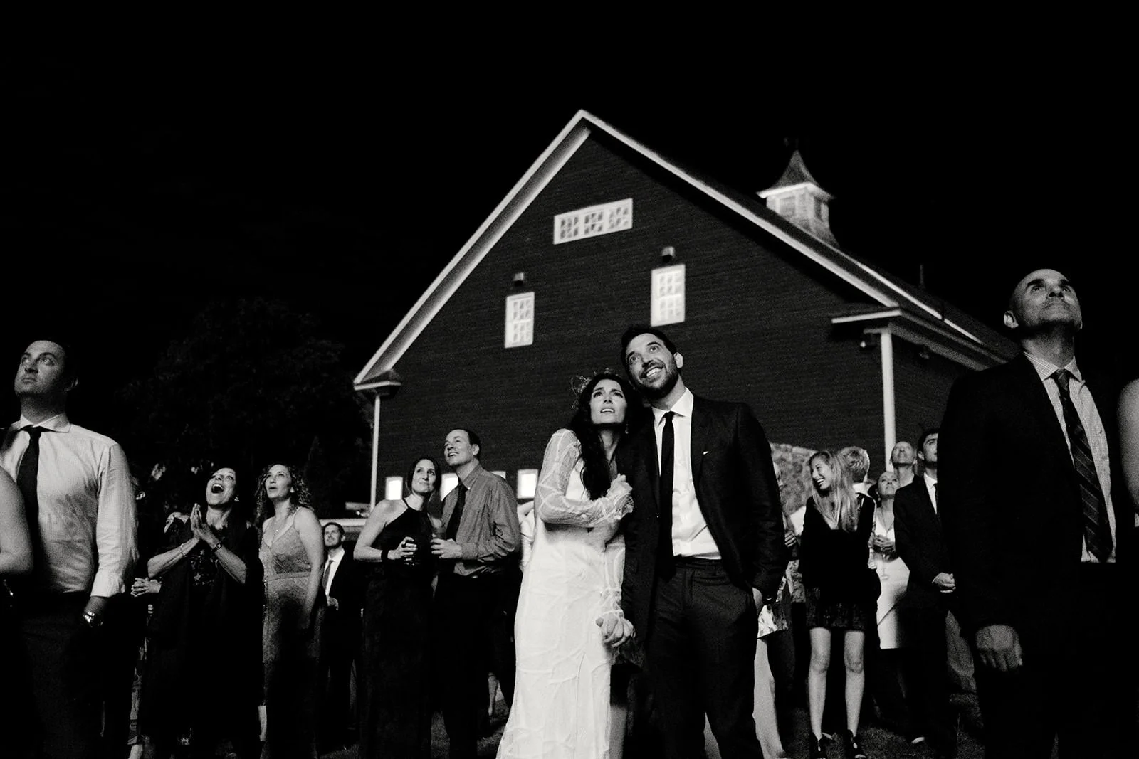 Black and white photo of a wedding reception outdoors at night, with a group of guests looking up at the sky, including a bride and groom standing close together in the center.