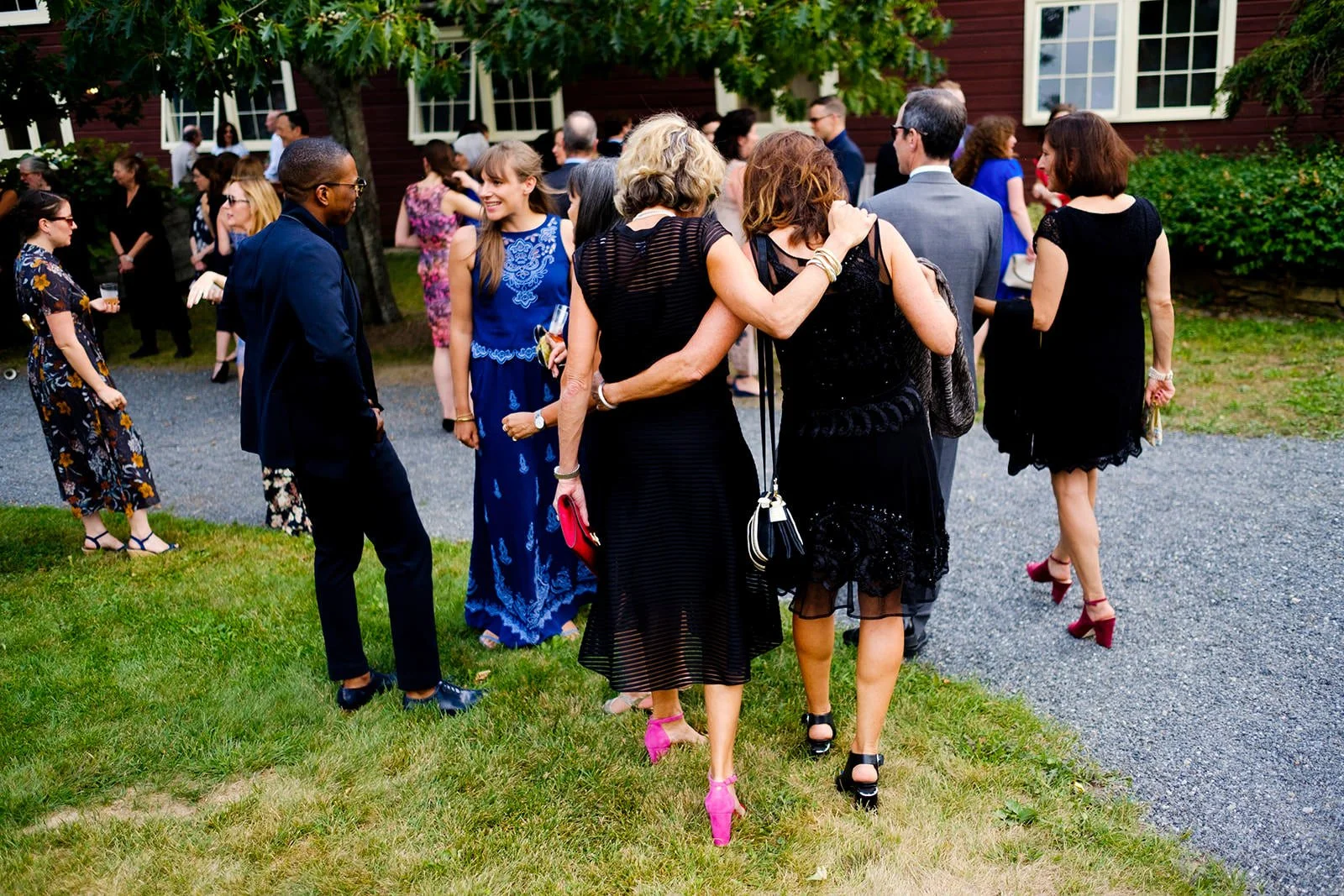 Group of people dressed in formal attire gathered outdoors on a lawn and gravel area at a social event with trees and a red building in the background.