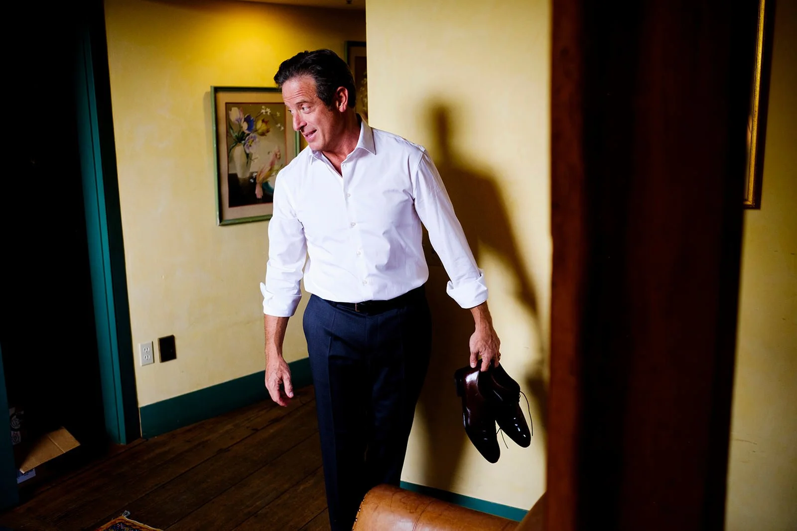 A man in a white shirt and dark pants is holding a pair of black shoes in his right hand, walking in a room with yellow walls decorated with framed artwork.