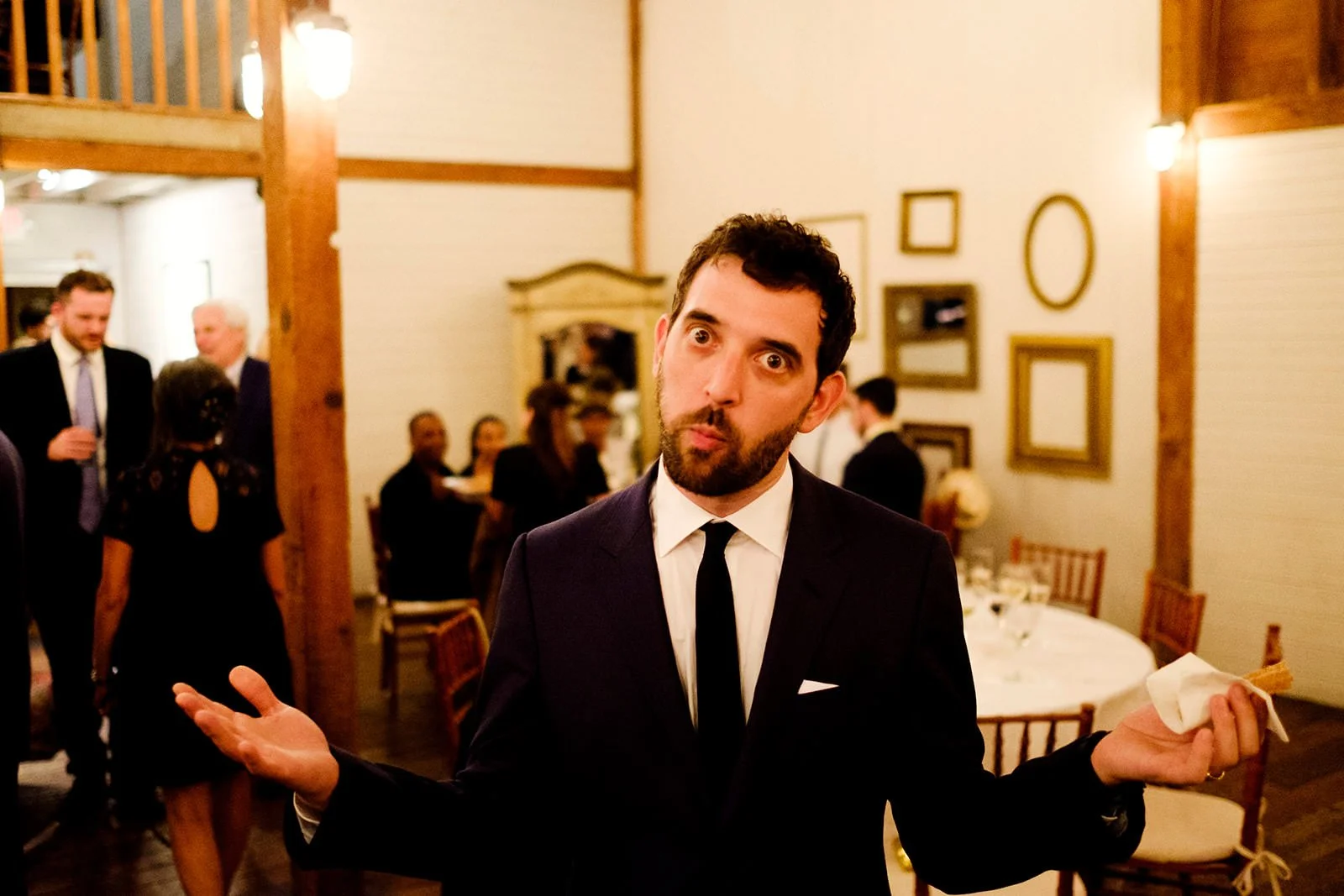 Man with dark hair and beard wearing a black suit and tie, making a humorous shrugging gesture with arms outstretched at a formal gathering in a decorated room with framed pictures on the wall.