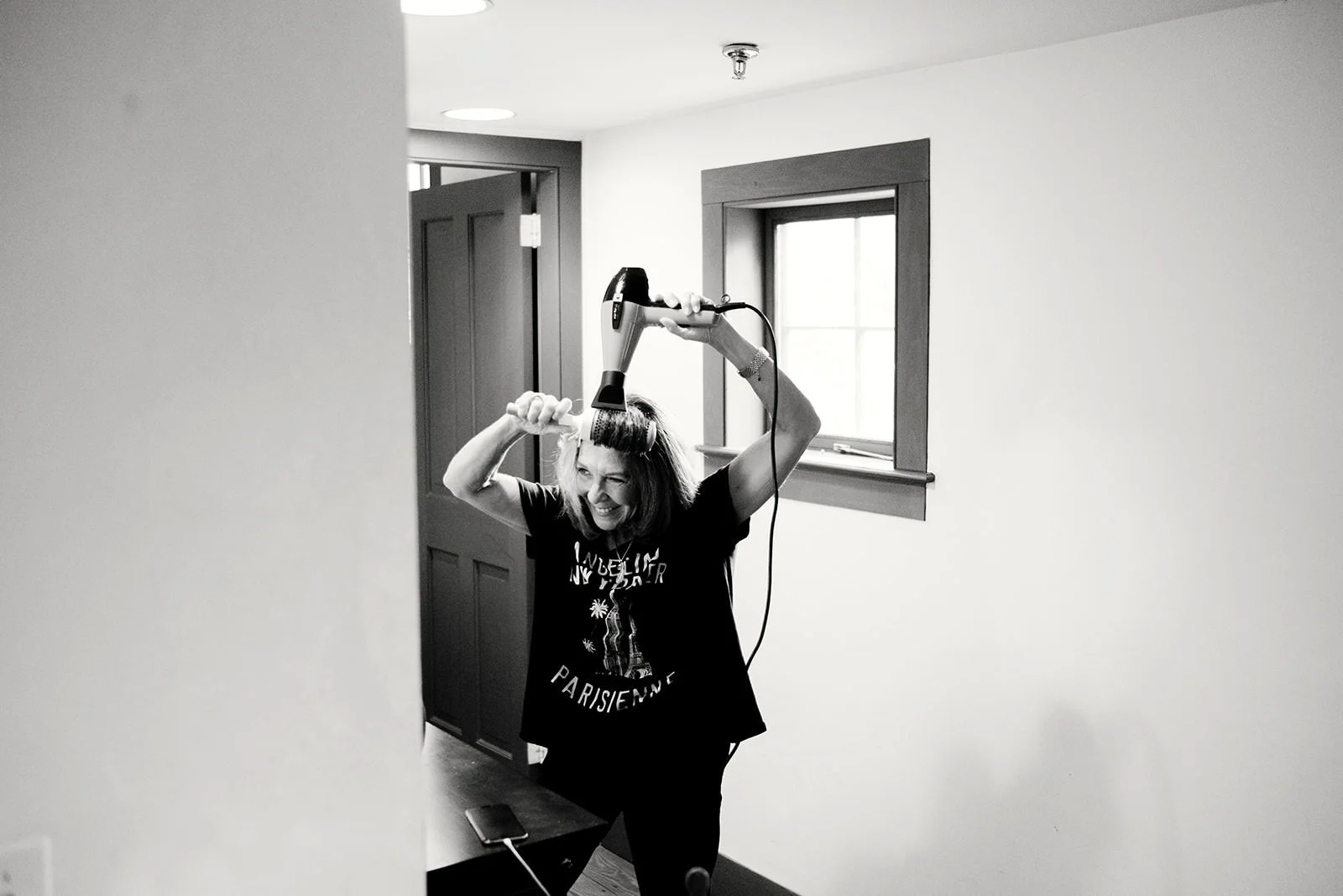 Woman with shoulder-length hair using a hairdryer on her head inside a room with a window and door, smiling and looking frustrated.
