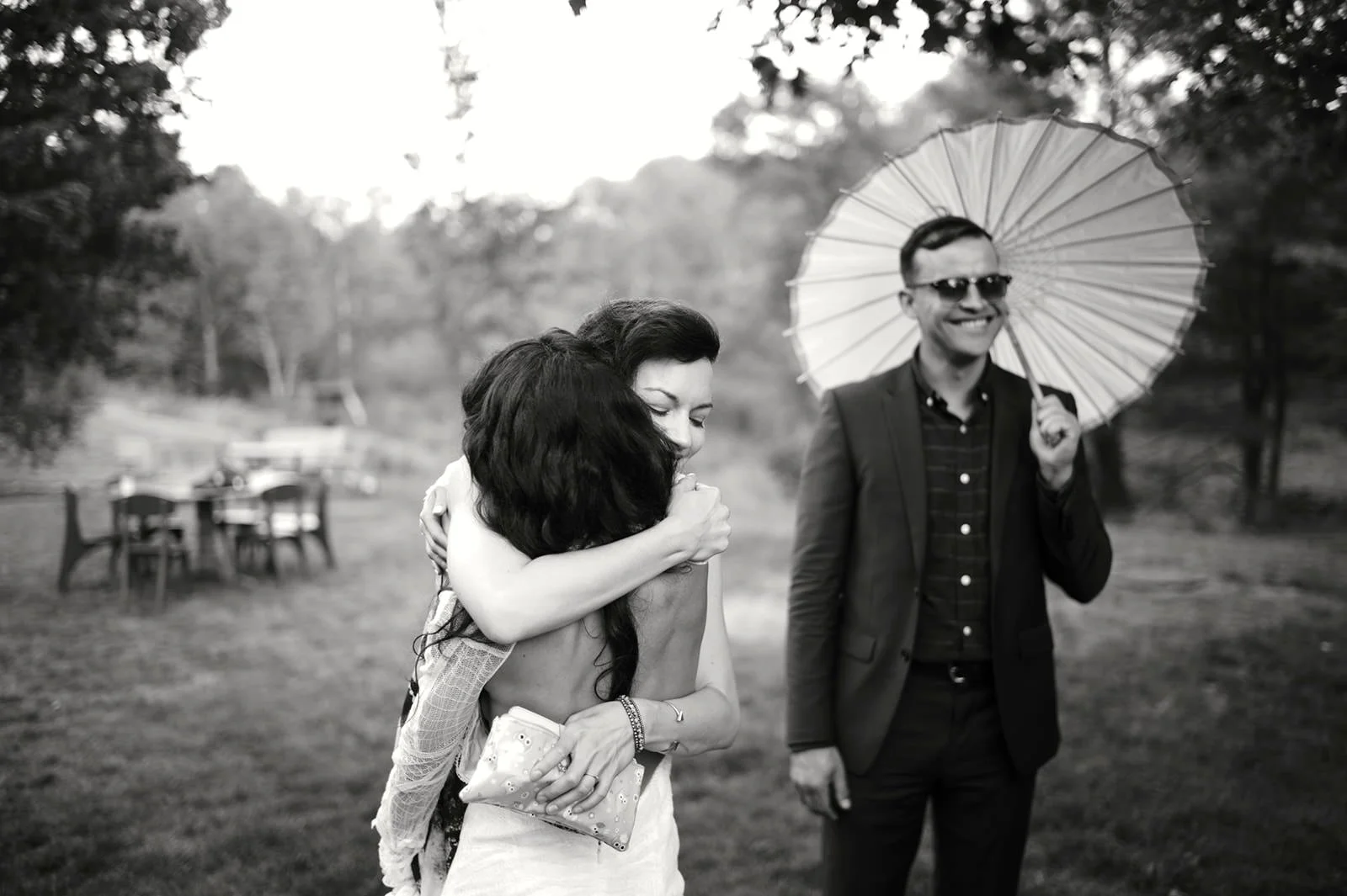 Black and white photo of two women hugging, with a man standing nearby holding an umbrella and smiling, in an outdoor park setting with trees and tables in the background.