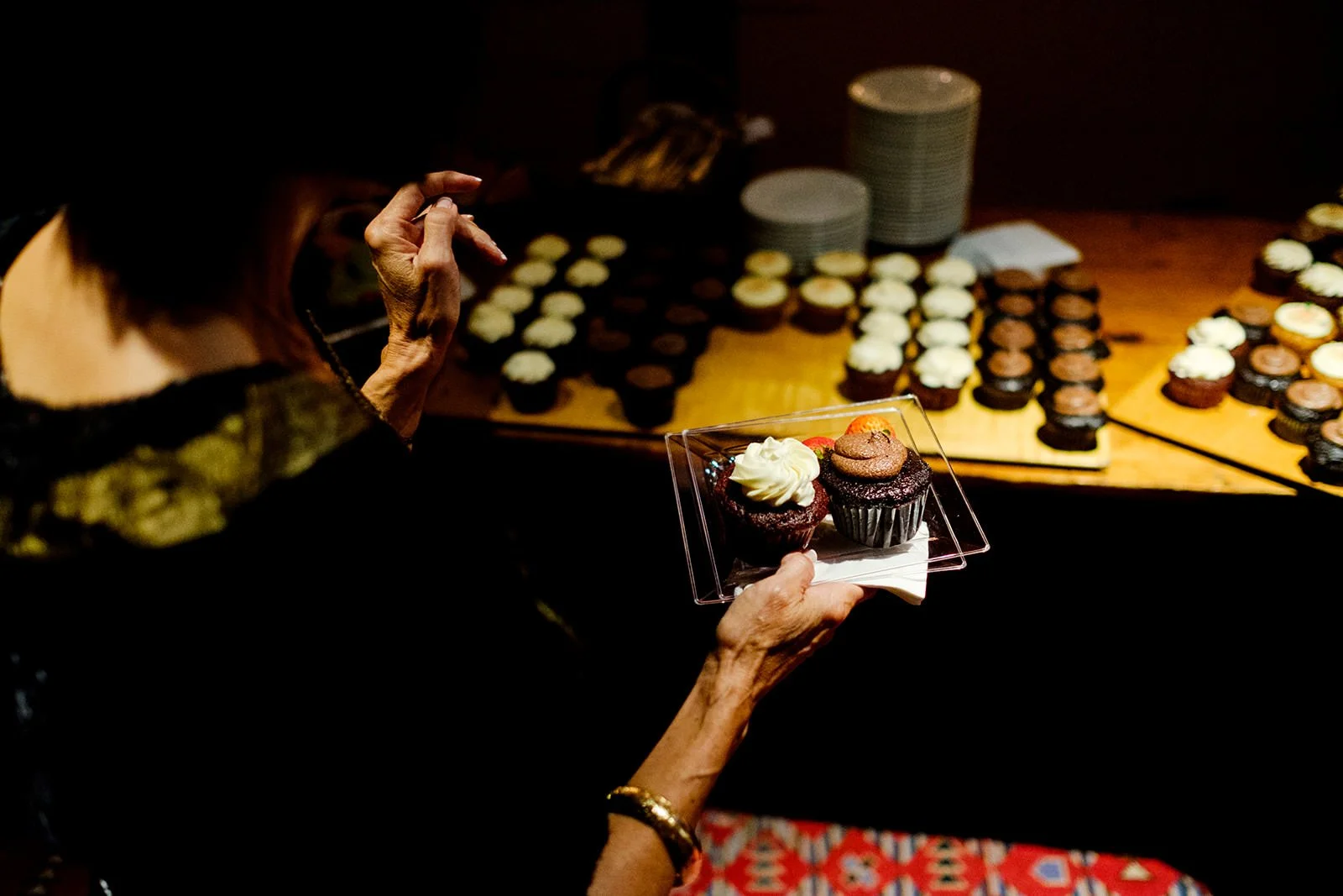 A person holding a plate with two decorated cupcakes in front of a table filled with more cupcakes.