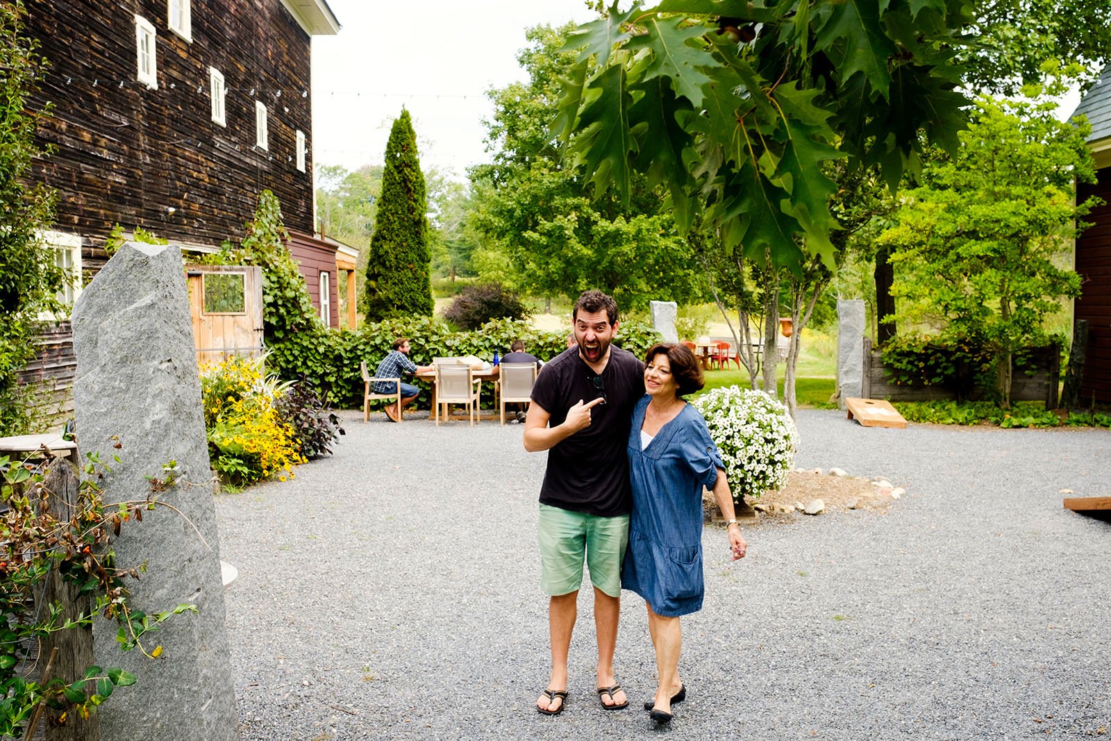 Two people, a man and a woman, are standing outdoors on a gravel surface, smiling and enjoying each other's company in a garden setting. The man is pointing at the woman.