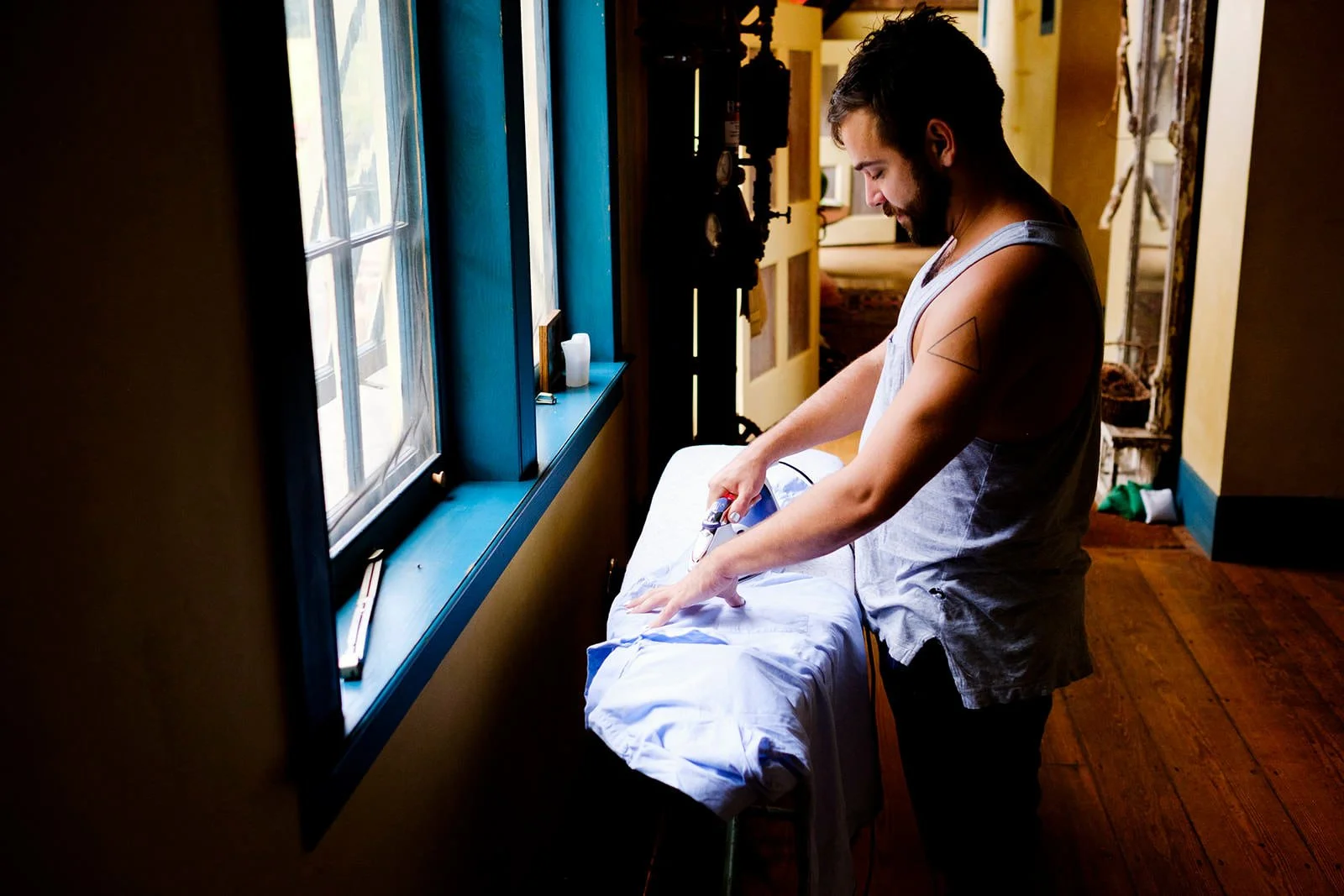 A man with a beard and tattoos on his arm is ironing a white shirt near a window in a room with yellow walls and wooden floors.