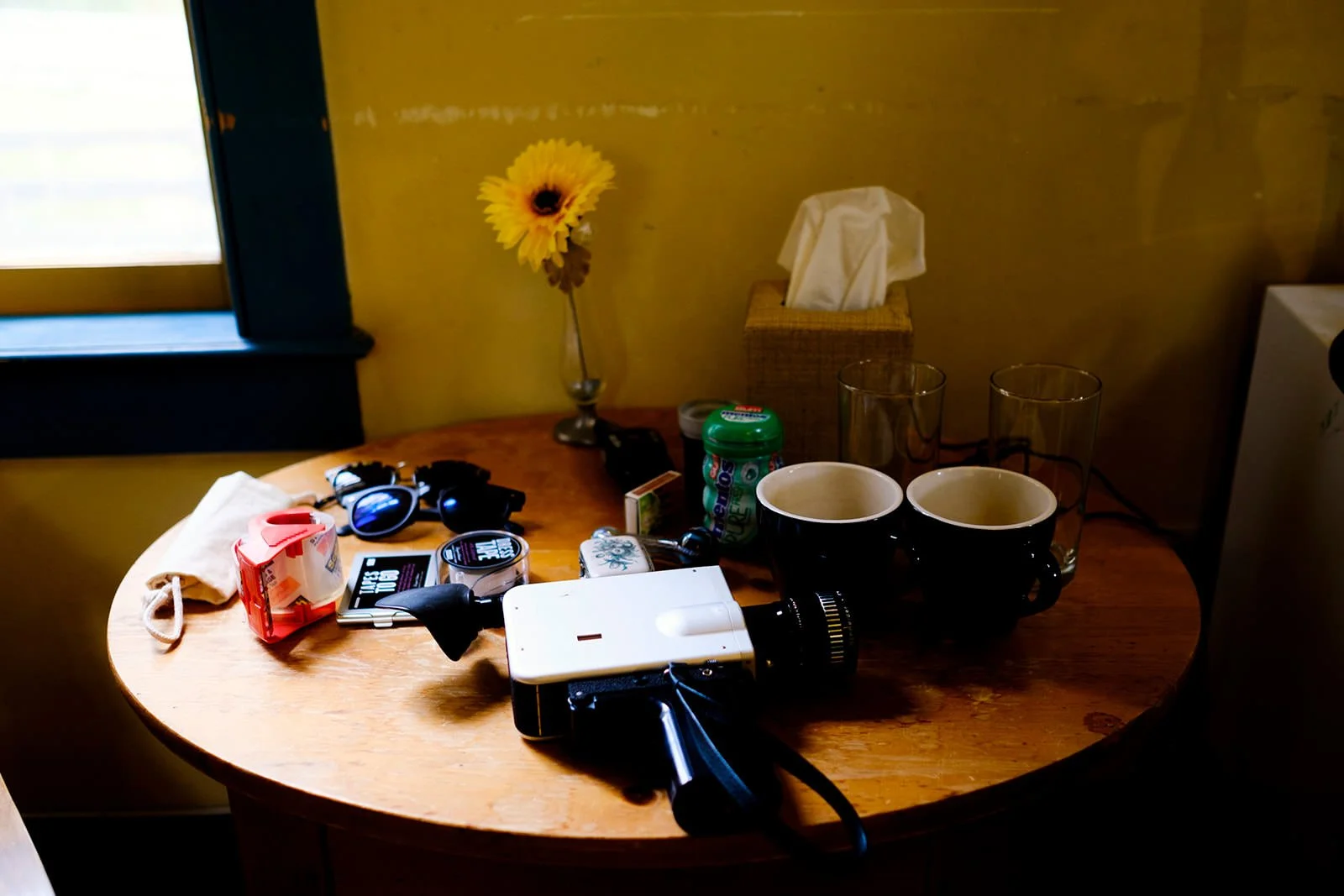 Round wooden table with various items: a camera, a pair of 3D glasses, a white smartphone, a small container, a bag of sugar, two black mugs, three drinking glasses, a green bottle, a tissue box, and a yellow flower in a vase, located near a window w