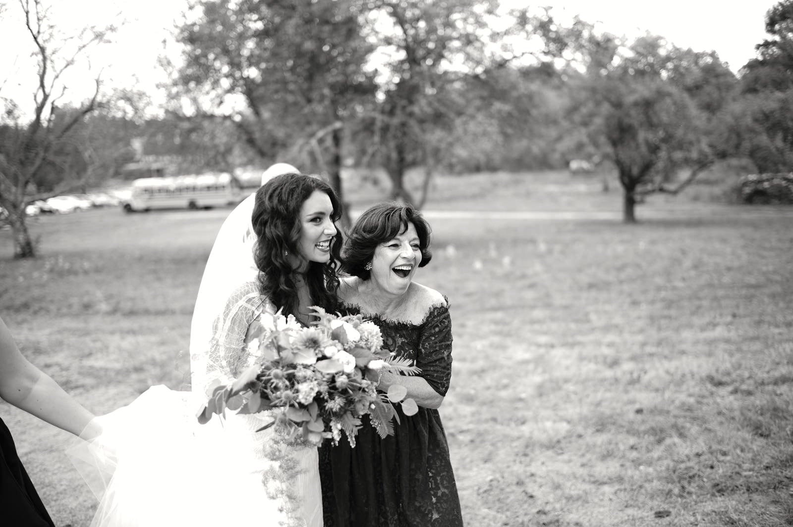 A bride in a wedding dress holding a bouquet of flowers, smiling, walking outdoors with a woman in a black dress, a veil partially visible, trees in the background.
