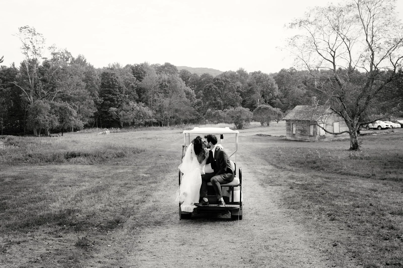 A black and white photo of a newlywed couple kissing on a small golf cart in a rural landscape with trees, a small wooden shed, and parked cars in the background.