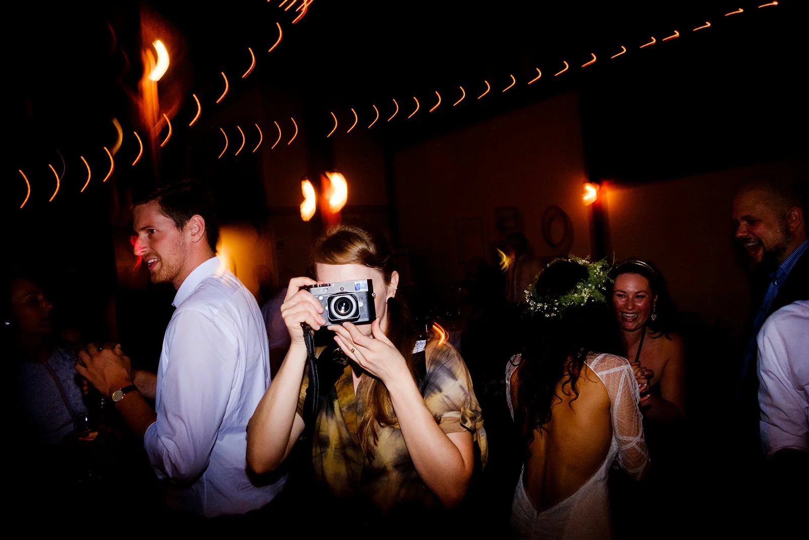 People enjoying a nighttime celebration or party indoors, with a woman taking a photograph and others socializing, decorated with string lights.