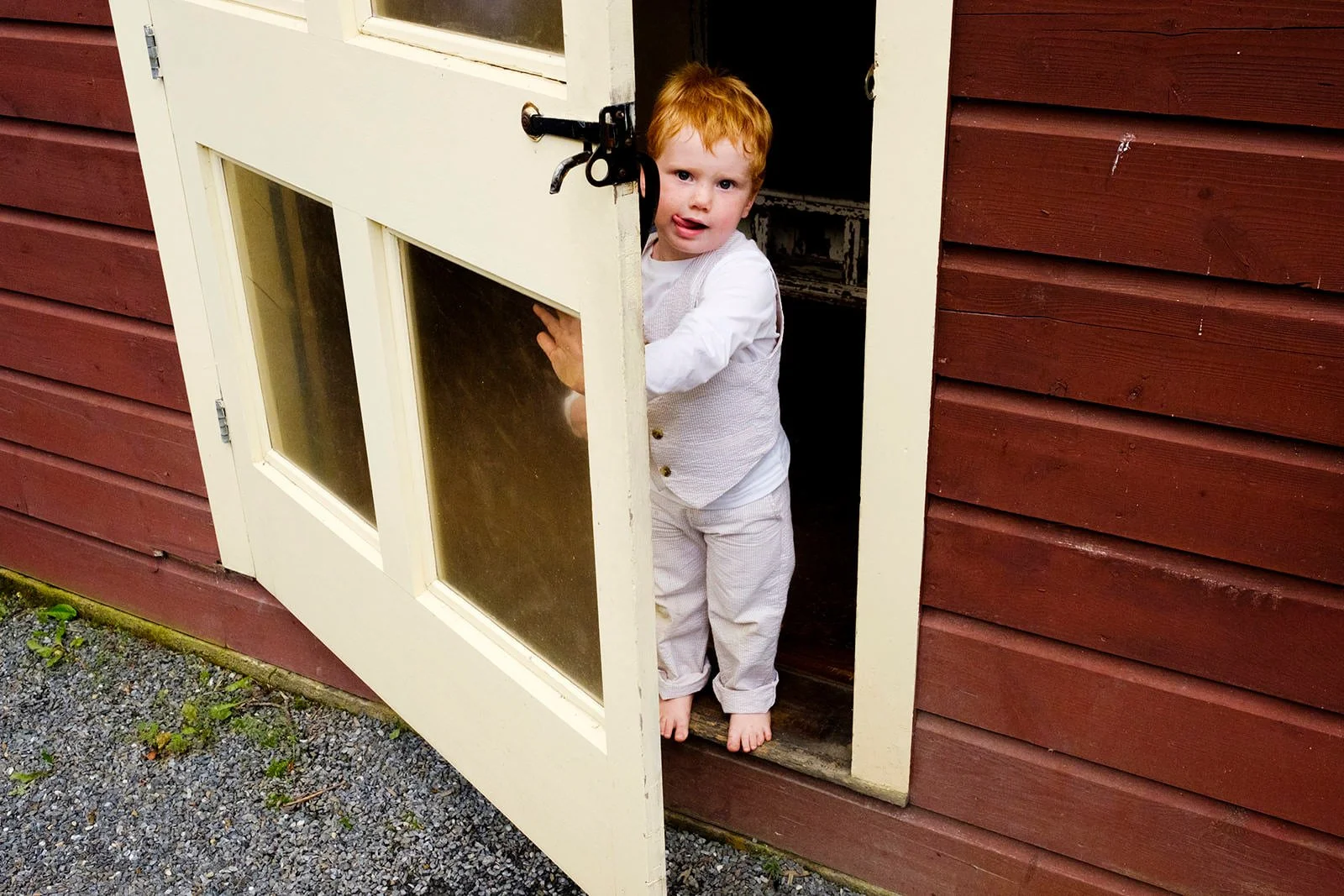 A young boy with red hair and white clothing standing barefoot at the door of a small wooden shed or playhouse, holding the door open and looking outside.