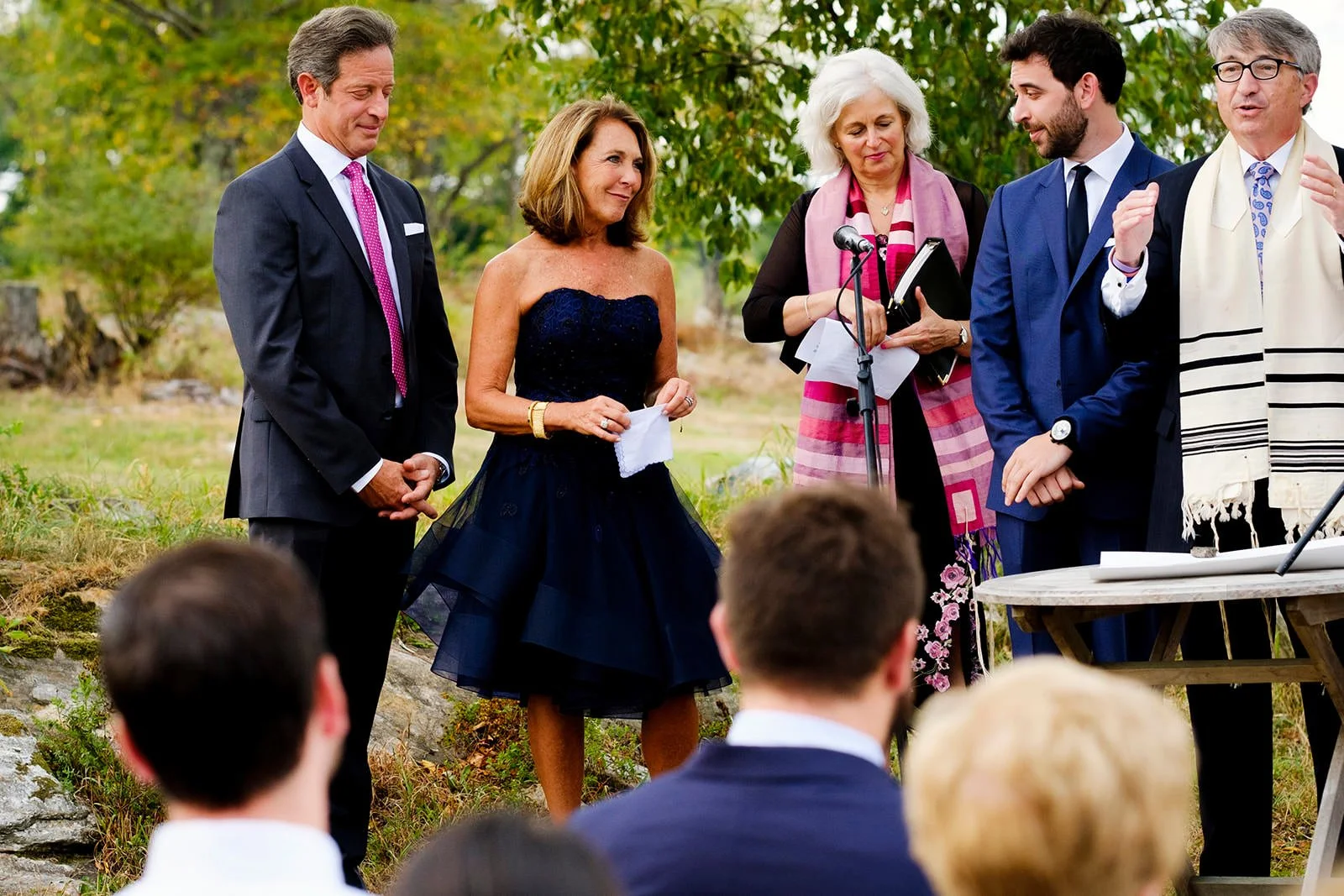 People gathered outdoors for a wedding ceremony, with a woman in a navy dress and a man in a suit standing in front of a microphone, while others listen and watch.