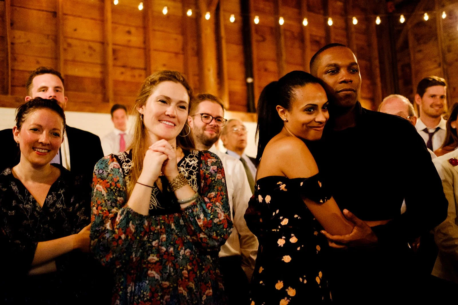 Group of people at a social gathering in a wooden venue, smiling, with string lights overhead, including a woman in a floral dress and a woman in a black off-shoulder dress leaning into a man dressed in black.