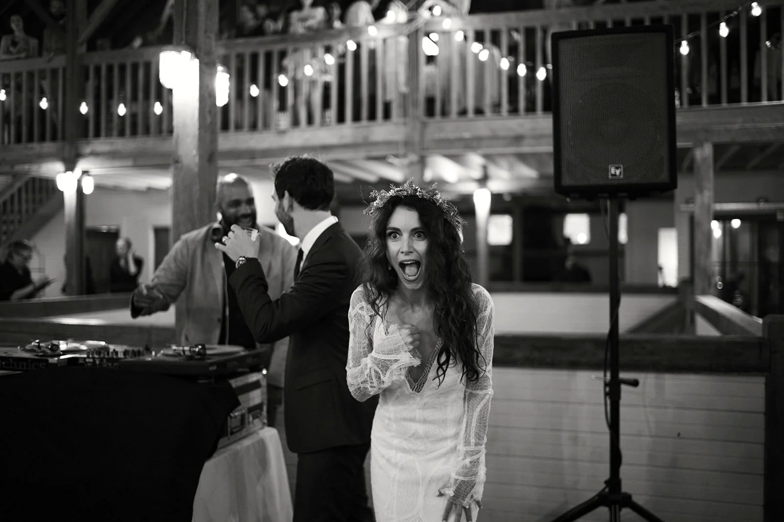 A woman in a white lace dress with a flower crown is shouting or singing with excitement at a party, while two men in suits are talking and smiling nearby. The setting appears to be an indoor venue with wooden decor and string lights.