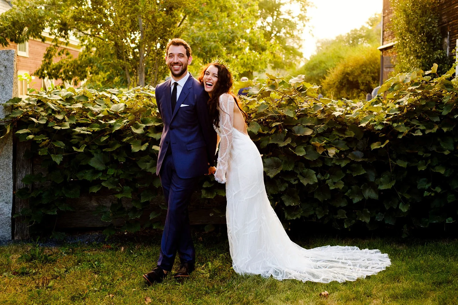 A happy couple, the groom in a navy suit and the bride in a white wedding dress, holding hands and laughing outdoors during sunset, with green trees and houses in the background.