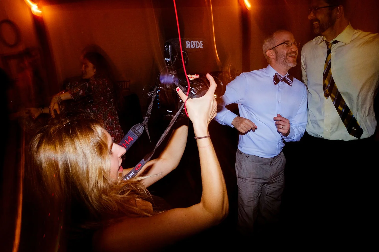 A woman with red hair is taking a photograph with a professional camera of two men with glasses who are smiling and dancing at a party or celebration.