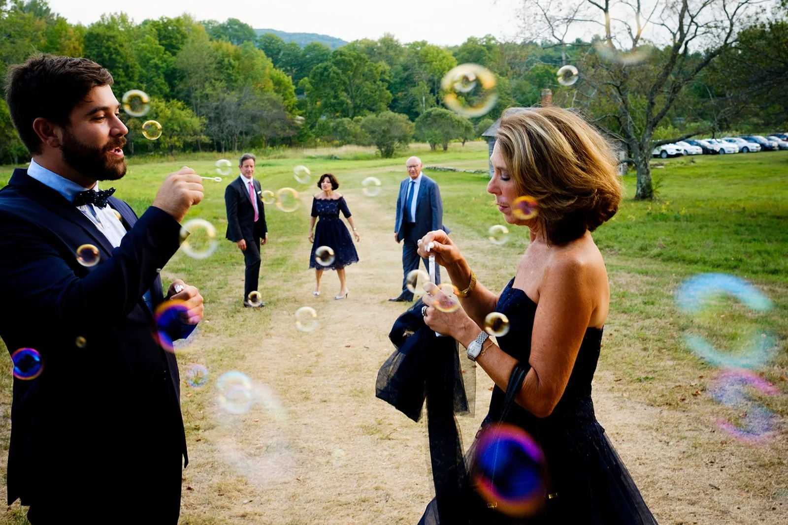 A group of people outdoors on a grassy path, with a man blowing bubbles and a woman in a dark dress preparing a bubble wand, while three others walk behind them in the background.