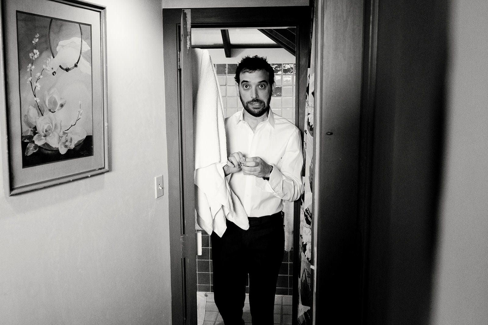 A man with dark hair and a beard, dressed in a white shirt and dark pants, standing in a narrow bathroom doorway holding a towel and a small item in his hands, with a surprised or curious expression on his face.