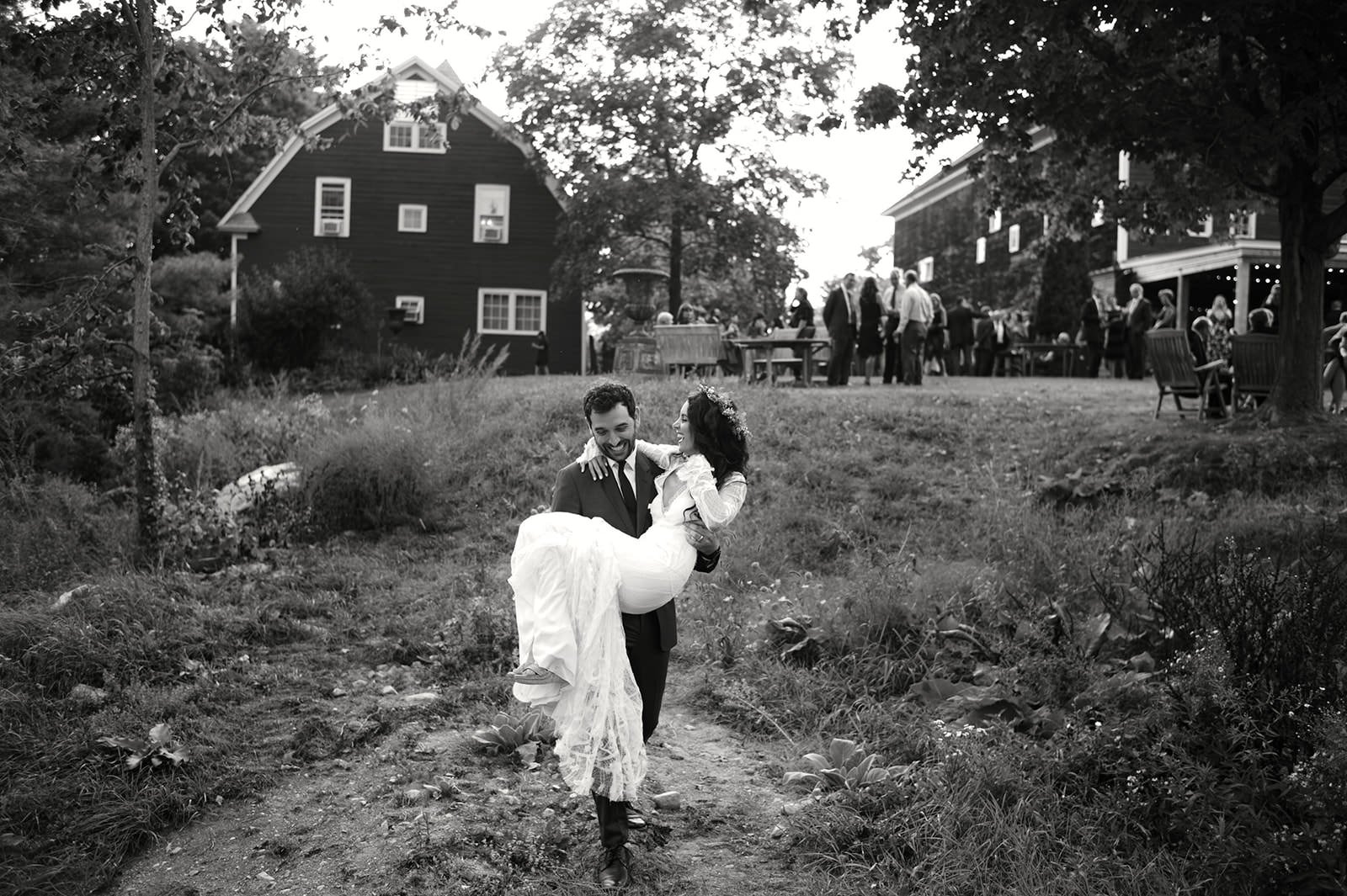A joyful wedding couple with the groom carrying the bride in a rustic outdoor setting, with guests in the background at a gathering
