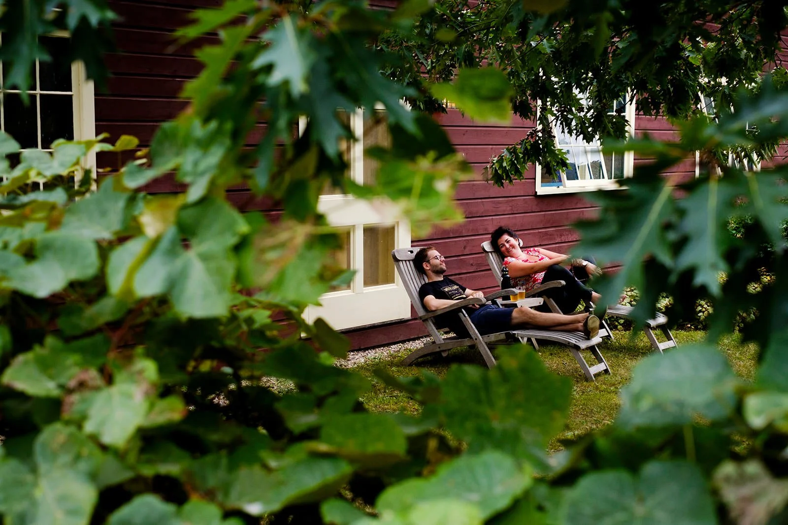 Two people relaxing on wooden lounge chairs in a garden with green foliage in the foreground and a red wooden house with windows in the background.