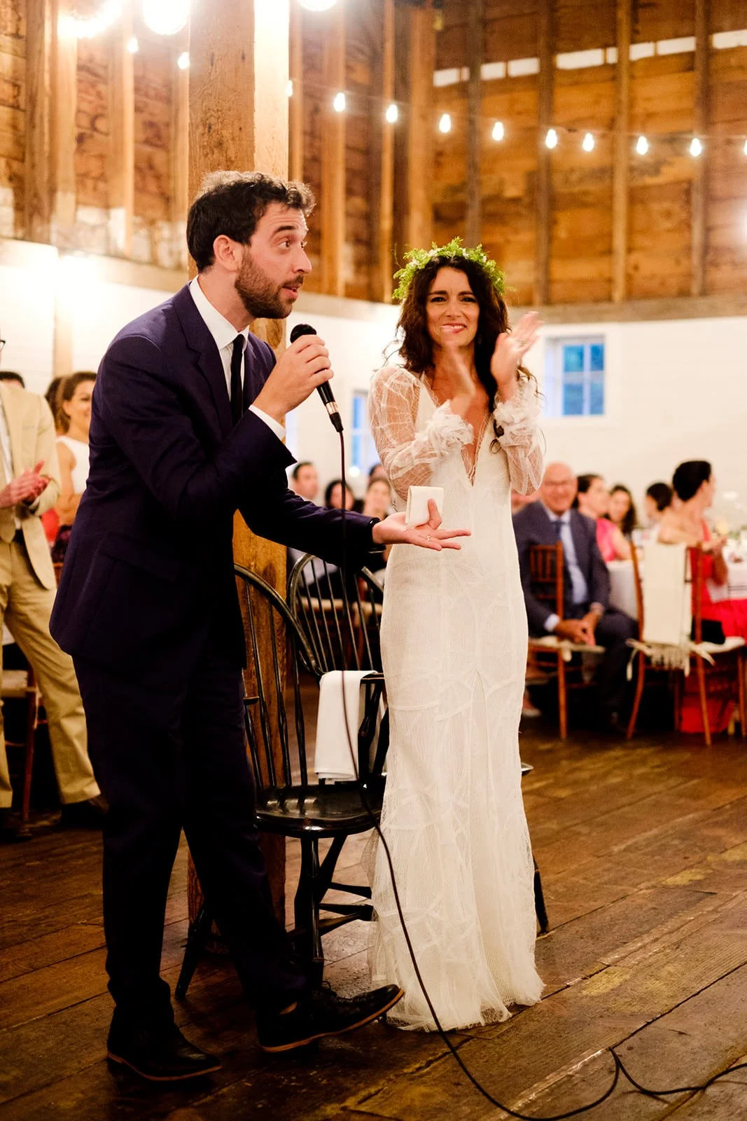 A man in a dark suit and a woman in a white wedding dress, with a flower crown, are standing inside a rustic barn, giving a speech at a wedding reception. The man is holding a microphone, and the woman is smiling and clapping. Guests are seated in th