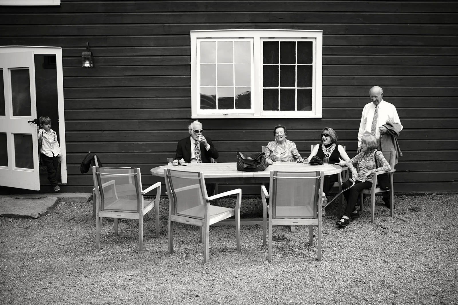 A group of five adults sitting and standing around a table outside a dark wooden building, with one child peeking out from the door, in black and white photo.
