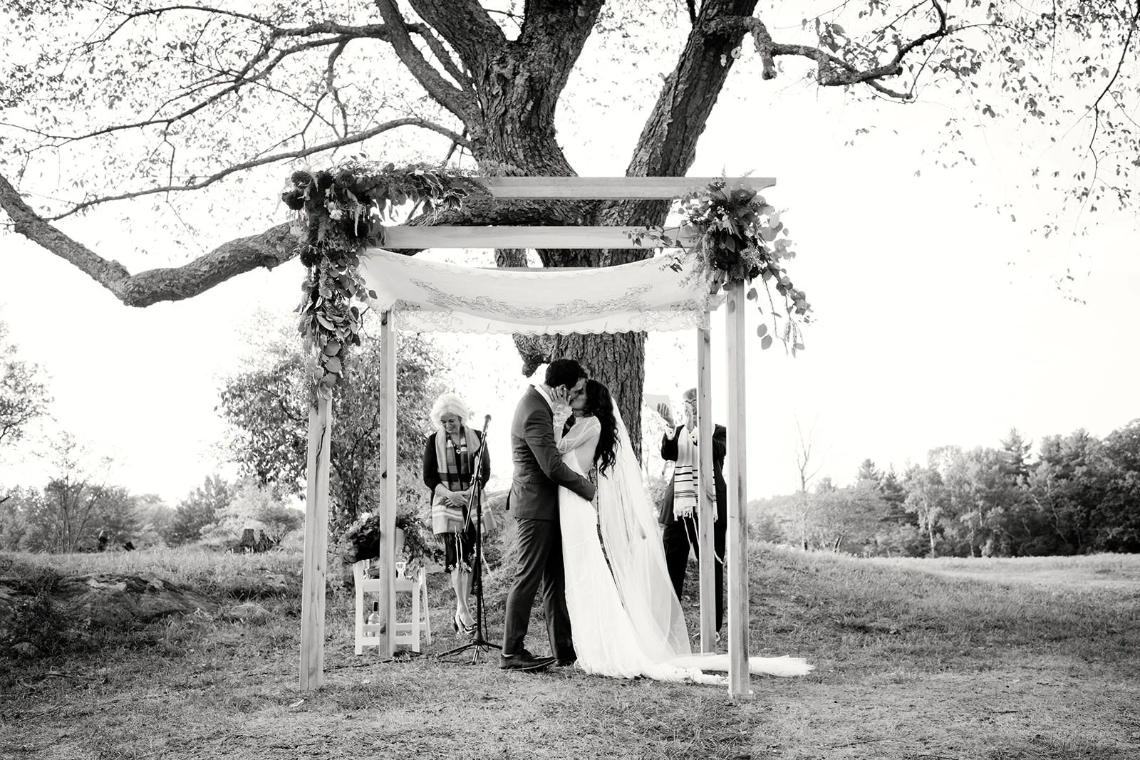 A black-and-white photo of a wedding ceremony outdoors. A couple is kissing under a decorated wooden arch, with a woman reading from a paper nearby. The ceremony is set in a grassy area with a large tree in the background.