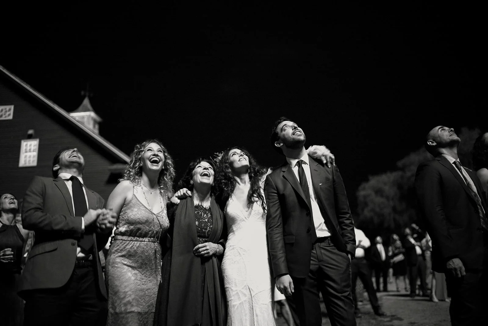 A group of people dressed in formal attire, looking up and smiling at an outdoor nighttime event with dark sky and building in the background.