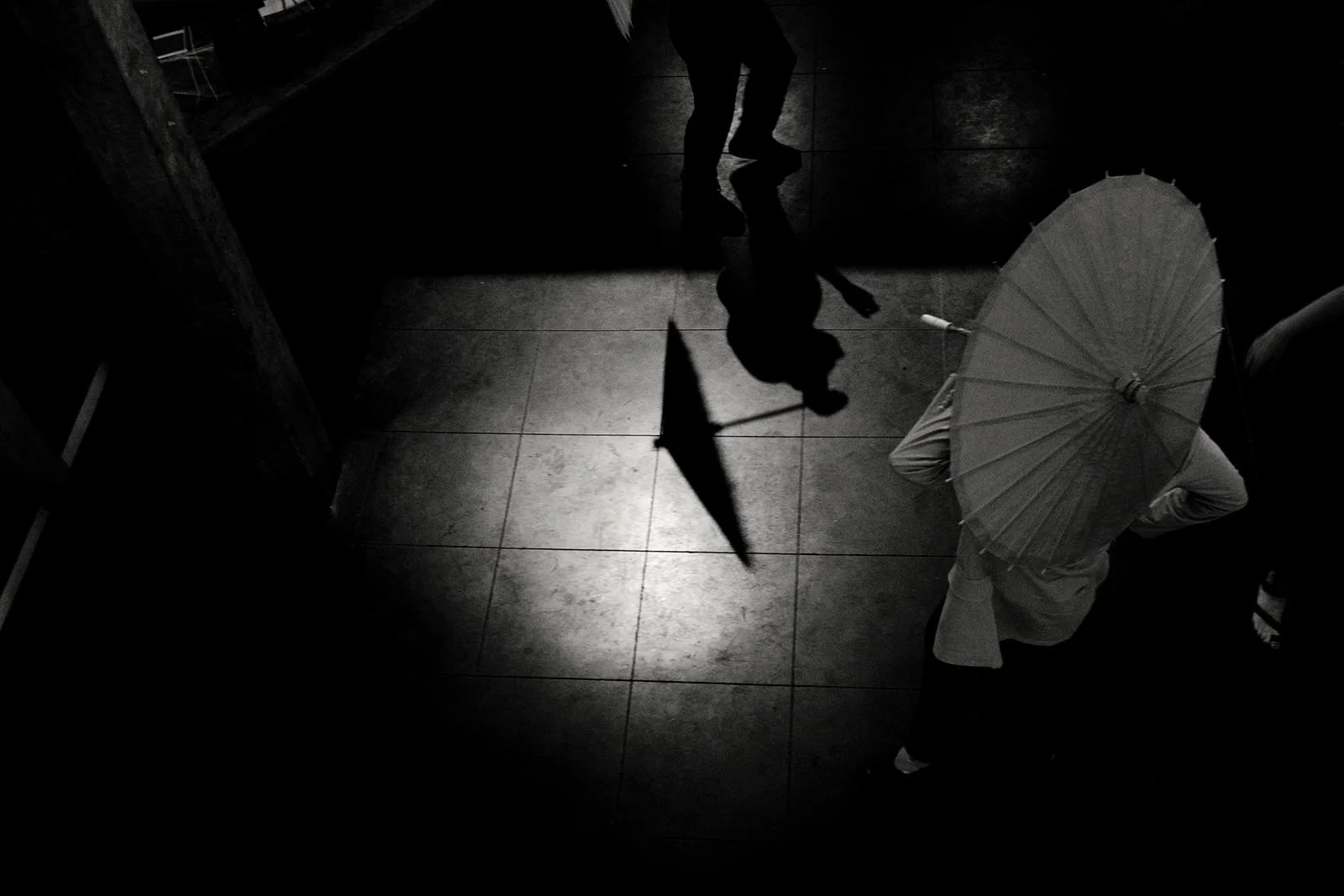 Silhouette of a person walking while holding an umbrella, with their reflection visible on the tiled floor, and another person in the background.