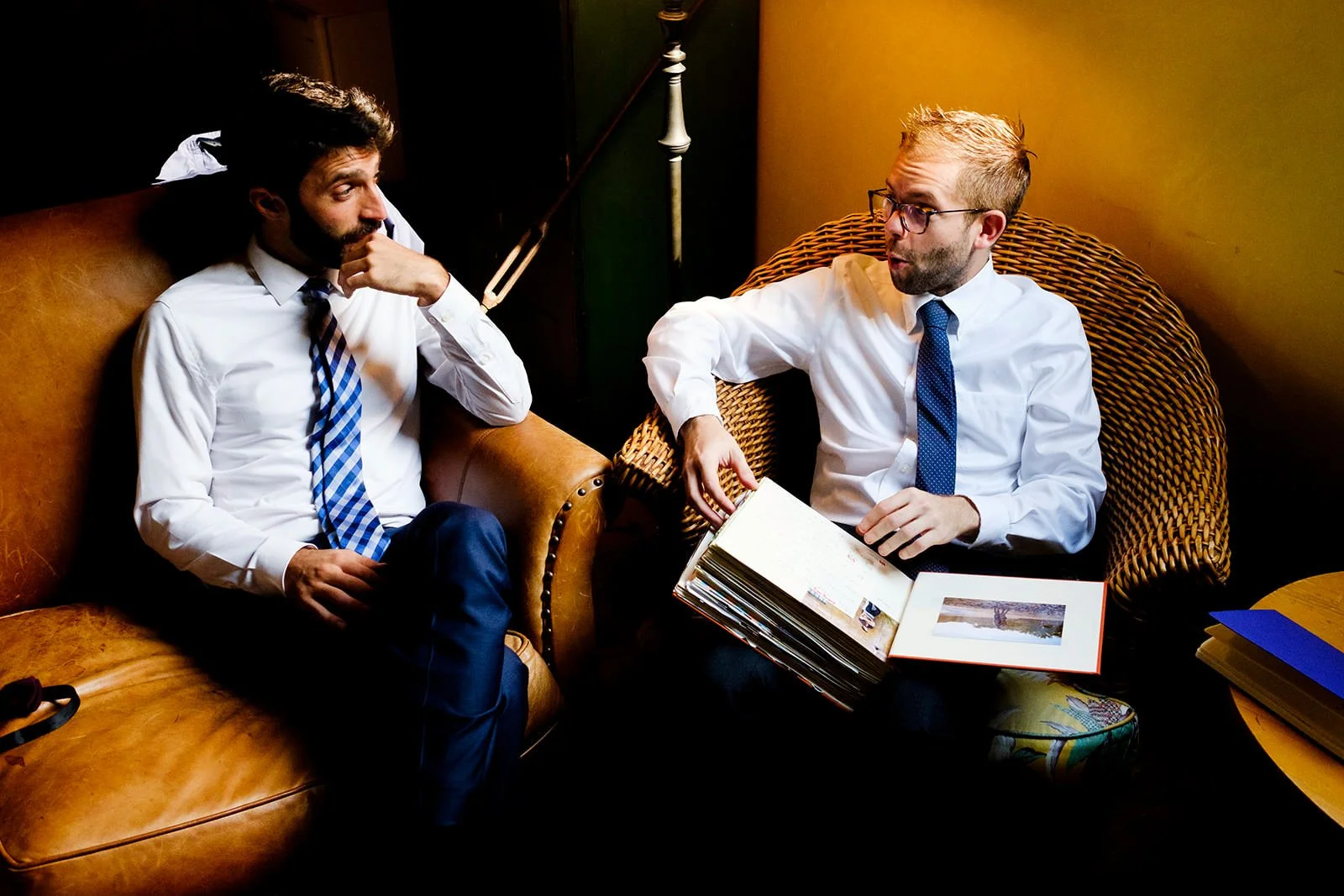 Two men are sitting in a room, one on a leather couch and the other on a wicker chair, engaged in conversation. The man on the right is holding a photo album open.