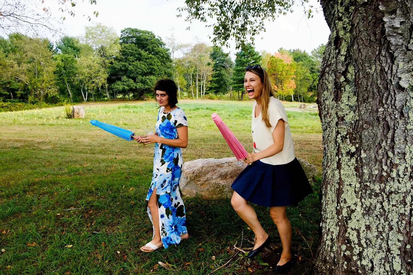 Two women outside near a large tree, one with a blue umbrella and the other with a pink umbrella, laughing and enjoying a playful moment.