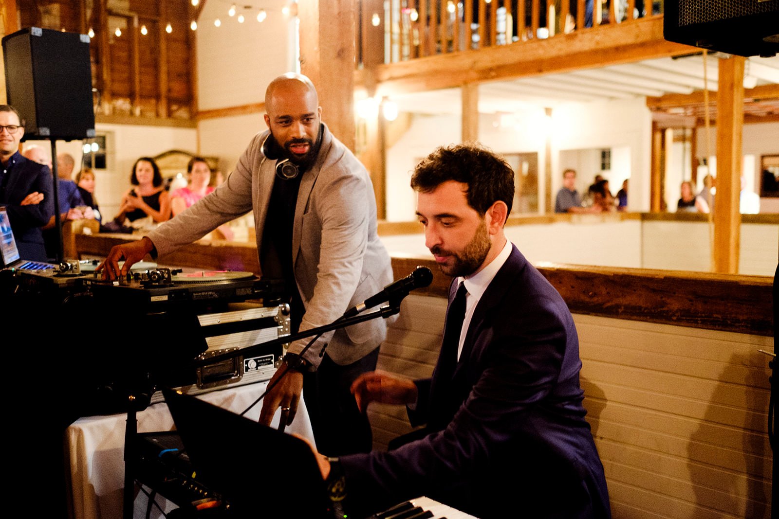 Two men, one with a headset and DJ equipment, the other playing a keyboard with a microphone, performing at a social event in a warmly lit wooden venue with guests in the background.