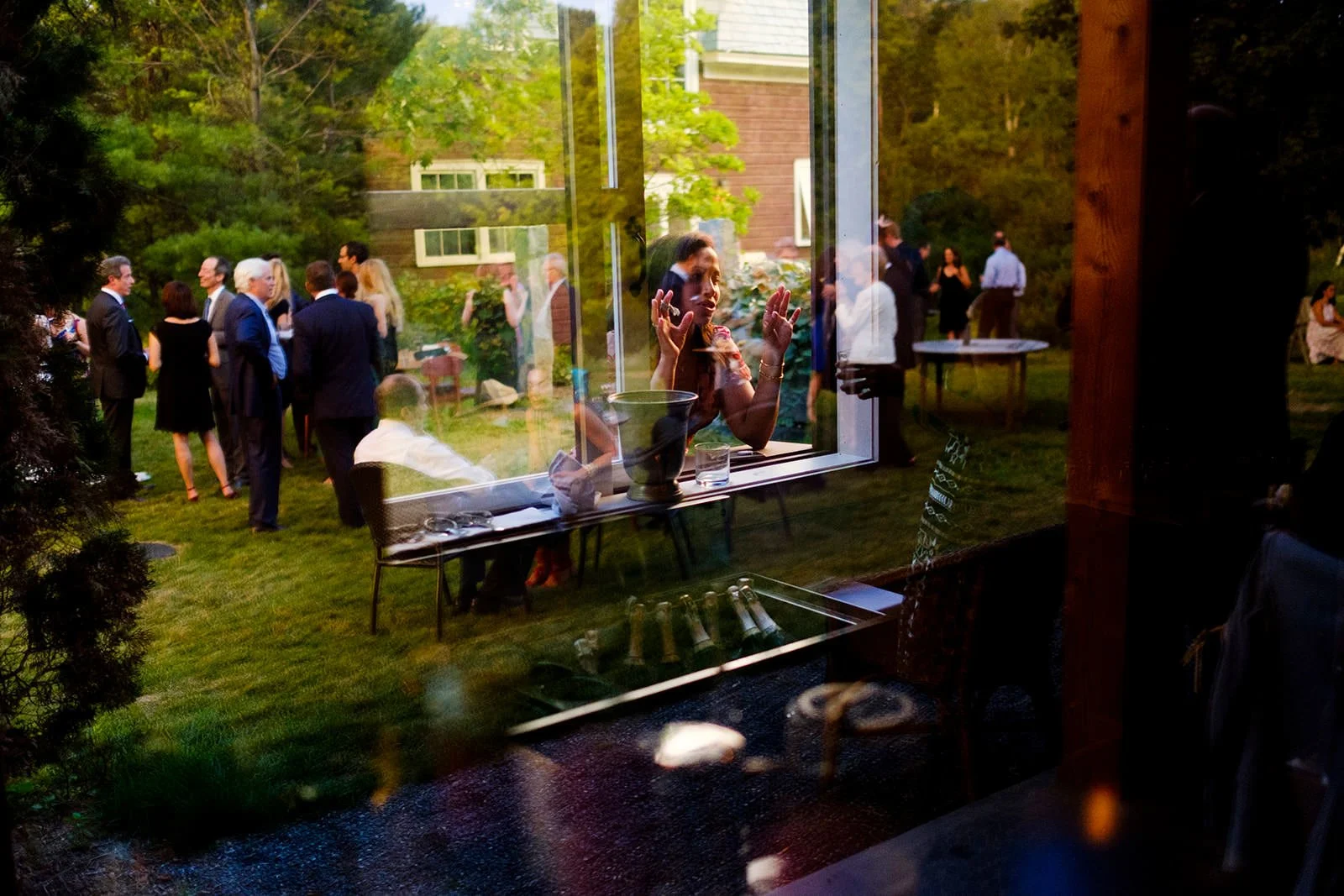 People gathered outside in a garden during a social event, viewed through a window with reflections and a woman inside reaching out to the window.