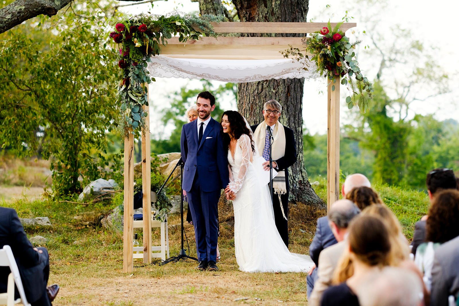 Wedding ceremony outdoors with a bride and groom holding hands, standing under a wooden arch decorated with flowers and greenery, surrounded by trees and guests seated in chairs.