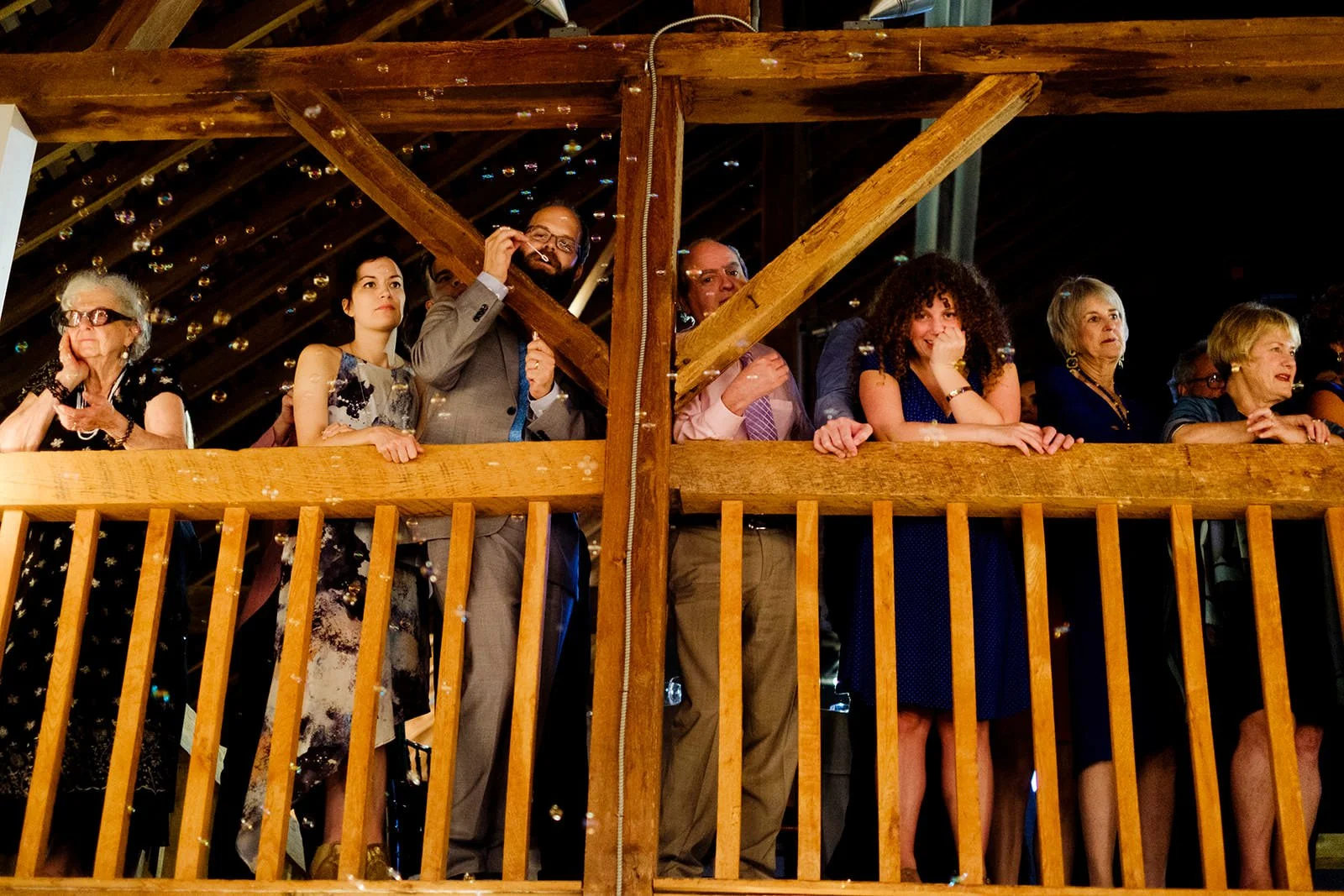 Group of people standing on a balcony watching an event, with some blowing bubbles and showing varied expressions.