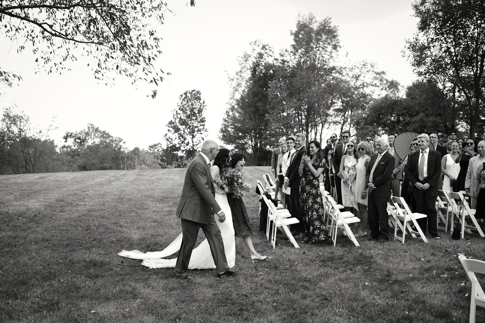 Black and white photo of a wedding ceremony outdoors. A bride and groom are walking down the aisle, smiling, while guests stand and watch. The guests are dressed in formal attire, with some holding umbrellas and sunglasses. The setting is on a grassy