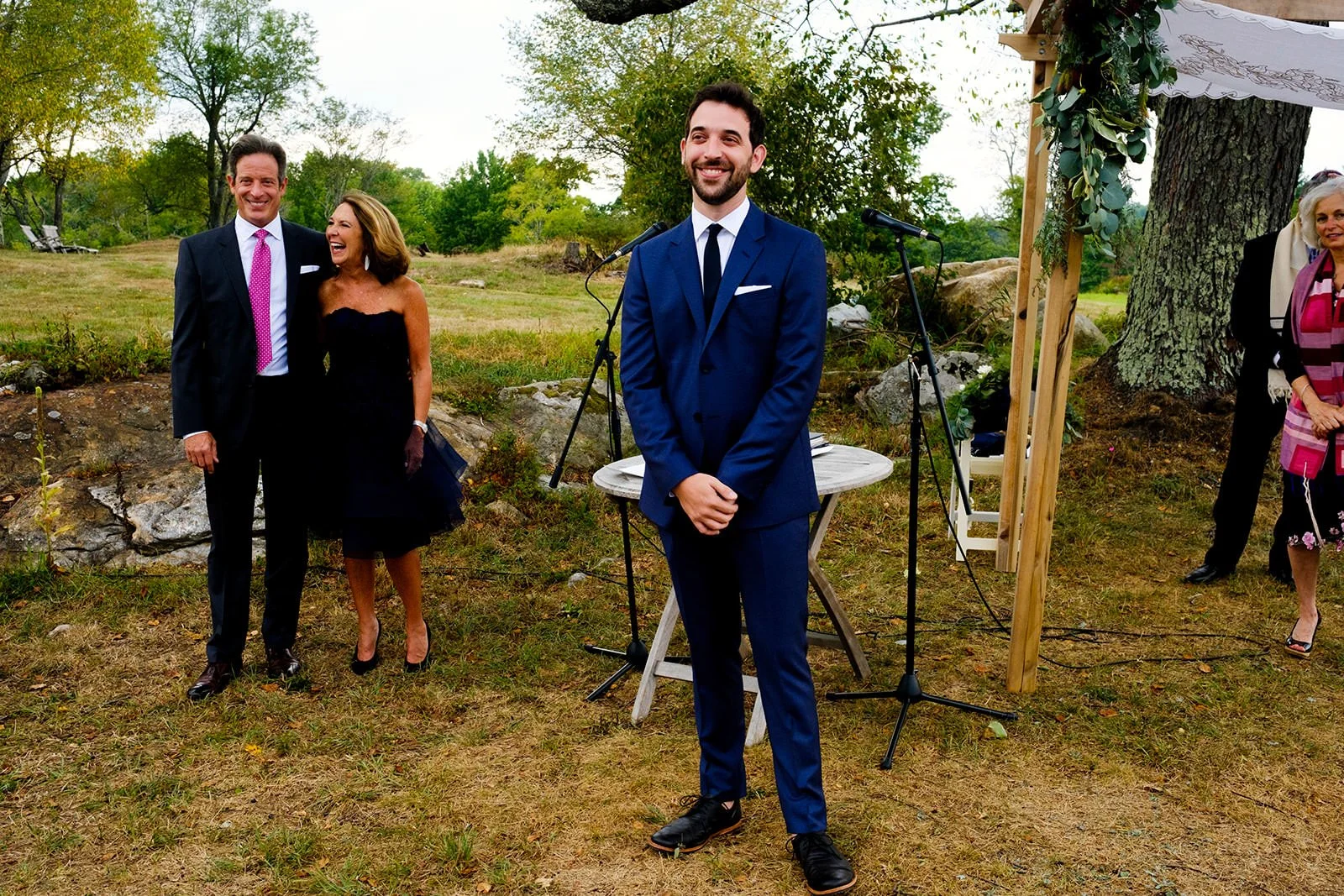 A man in a blue suit stands in front of an outdoor wedding altar, smiling with his hands clasped. Behind him, two people are laughing: a man in a dark suit and a woman in a black dress. To the right, a woman in a patterned dress is partially visible,