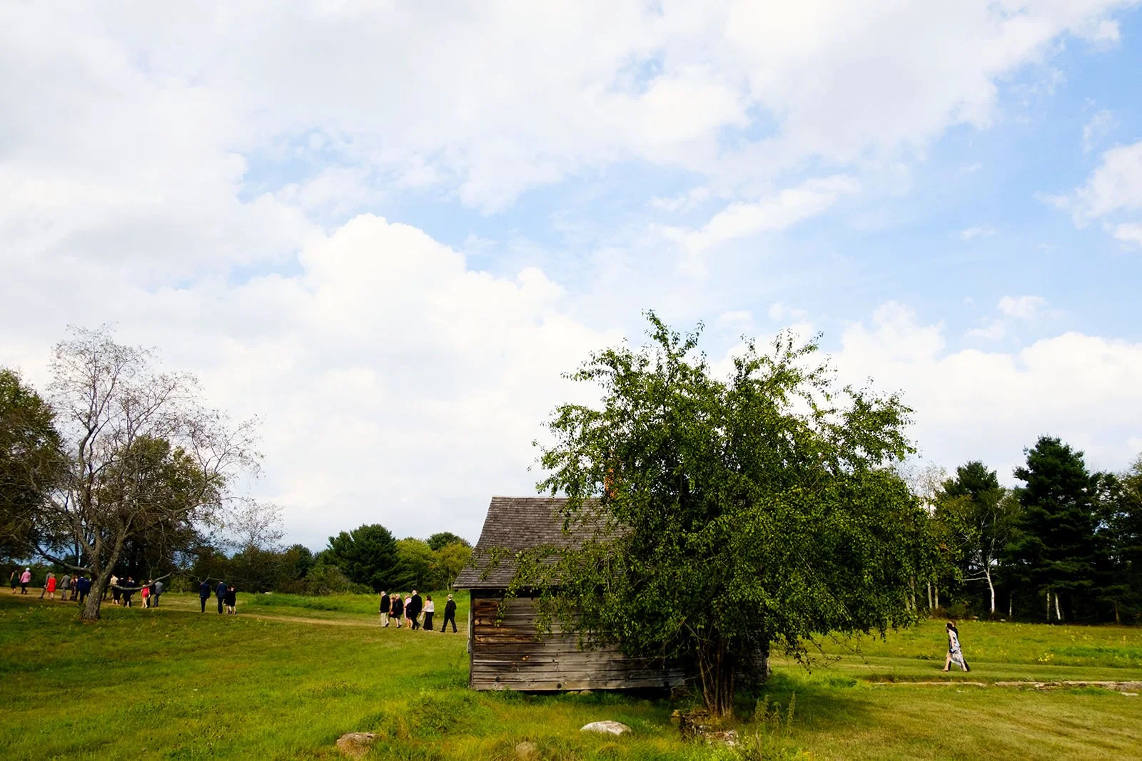 A rustic wooden barn with a shingled roof standing in a grassy field, partly covered by a leafy tree, with people walking in the distance under a partly cloudy sky.