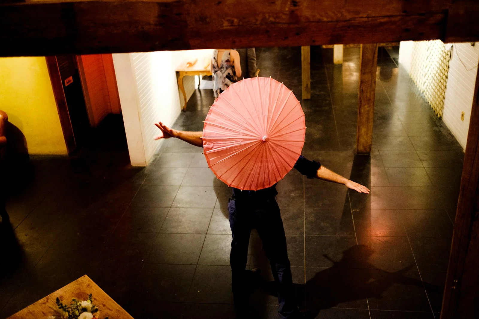 Person holding a pink paper umbrella with arms extended, standing in a dimly lit room with dark tiled floor, and wooden beams overhead.