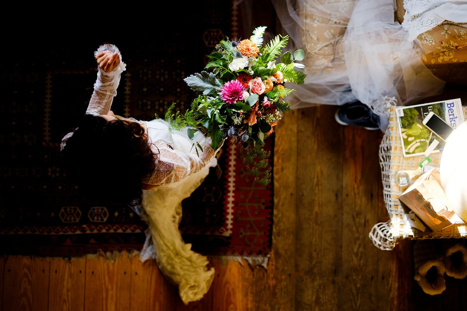 A woman with curly dark hair in lace clothing kneeling on a patterned carpet, holding a large bouquet of colorful flowers, with a fluffy white rug and a wooden floor nearby, and a wicker table loaded with magazines, a paper bag, and a smartphone.